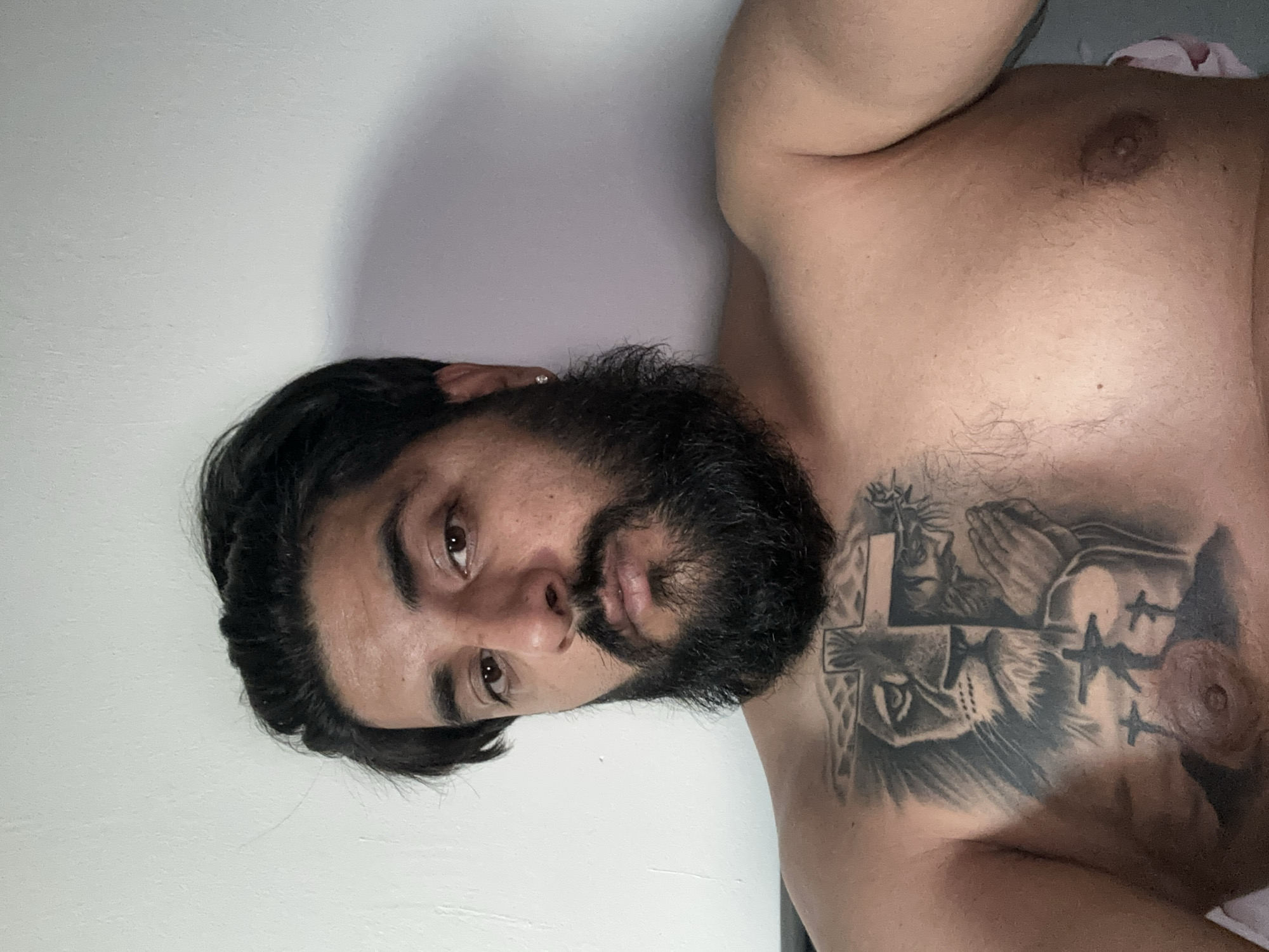 A man with dark hair and a beard is lying down, looking at the camera. He is shirtless, and has a tattoo on his chest. The tattoo depicts a lion's head, Jesus with praying hands, and three crosses on a hill. The background is a plain wall. The image was taken indoors, likely in a bedroom, in Mainburg, Germany.