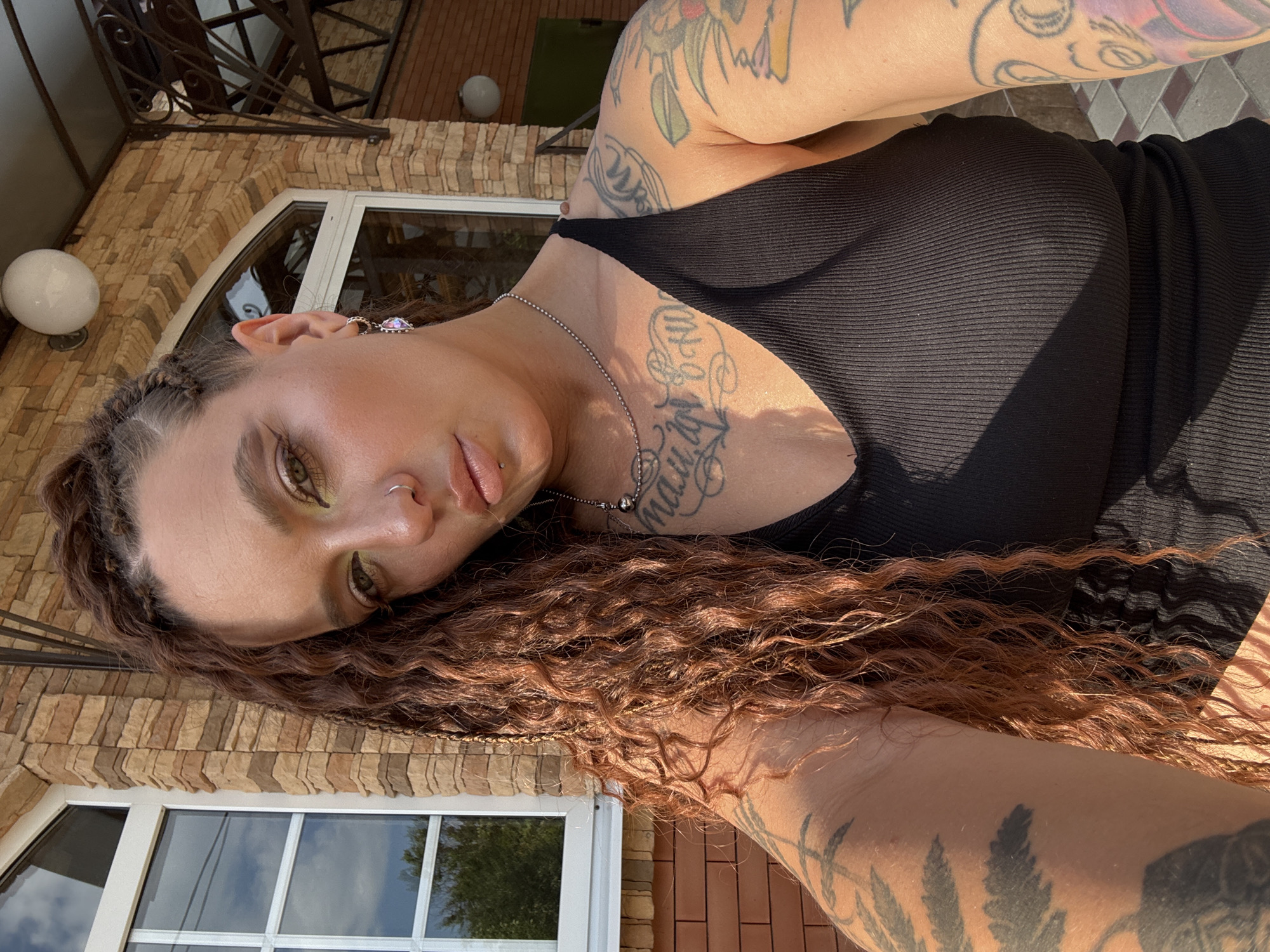 This is a vertically oriented, close-up photograph of a woman lying on her side, outdoors. She has long, wavy, reddish-brown hair with braids woven in. Her face is turned towards the camera, with her right eye in focus and her left eye partially obscured. She has noticeable makeup, including eyeshadow, and a nose ring. She is wearing a black, ribbed tank top and a silver necklace with a spherical pendant. Tattoos are visible on her arms and chest, some of which feature script. She is wearing a sparkly earring on her right ear.
The background shows a textured, light brown brick wall and a large window with reflections of the sky and trees. A white, globe-shaped light fixture is also visible on the wall. The lighting suggests it is daytime, with strong sunlight casting shadows, indicating fair weather. The overall impression is one of a casual, relaxed moment captured outdoors. There is no visible text in the image.