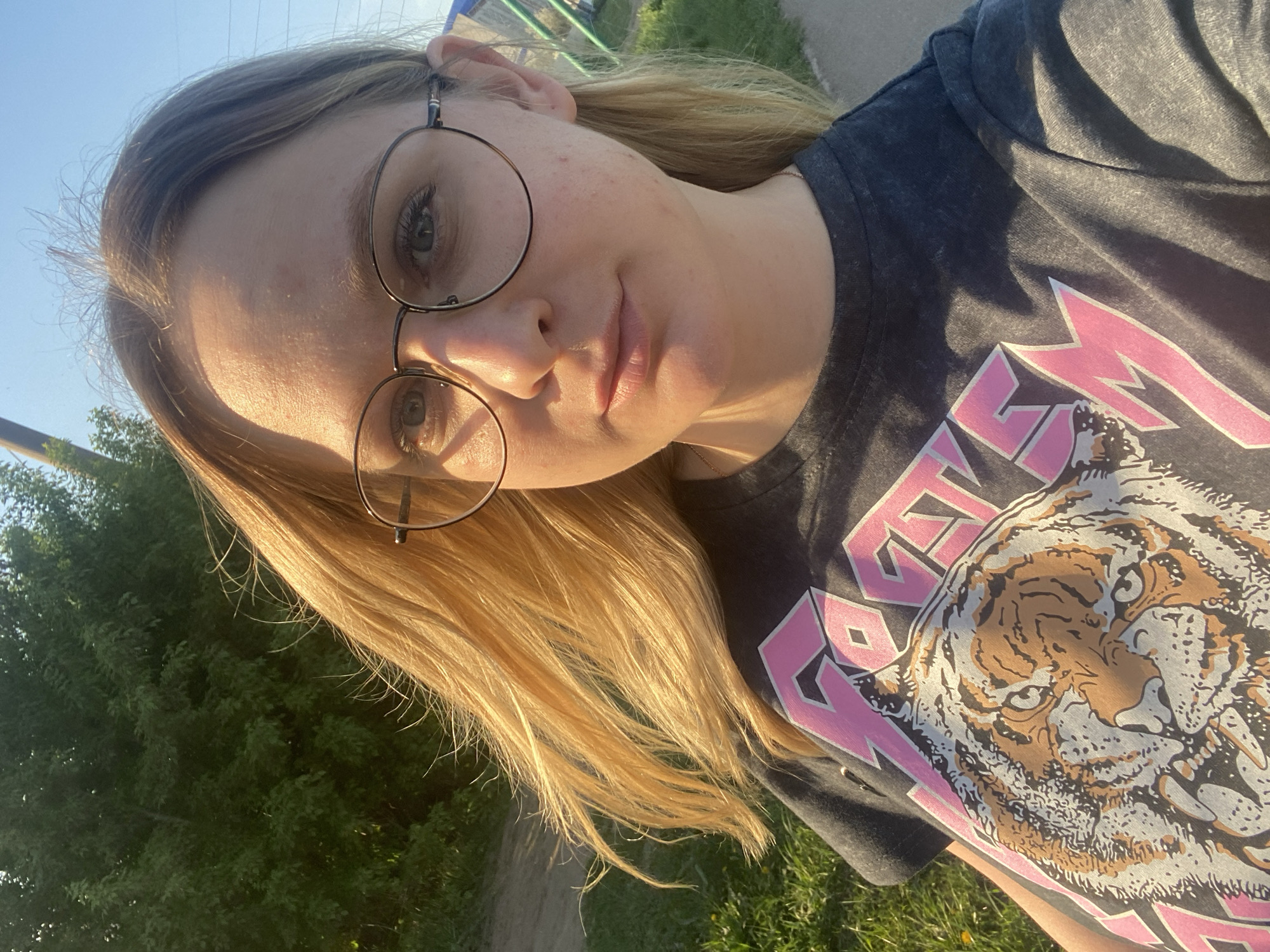 A person, likely female, is centered in a self-shot image. The subject has fair skin and light blonde hair, and wears round, wire-framed glasses. A gray t-shirt with a pink design and tiger print covers the upper torso. The background consists of green foliage, and the sky is visible in the upper left corner. The image appears to be taken outdoors, in the city of Unknown, Unknown.