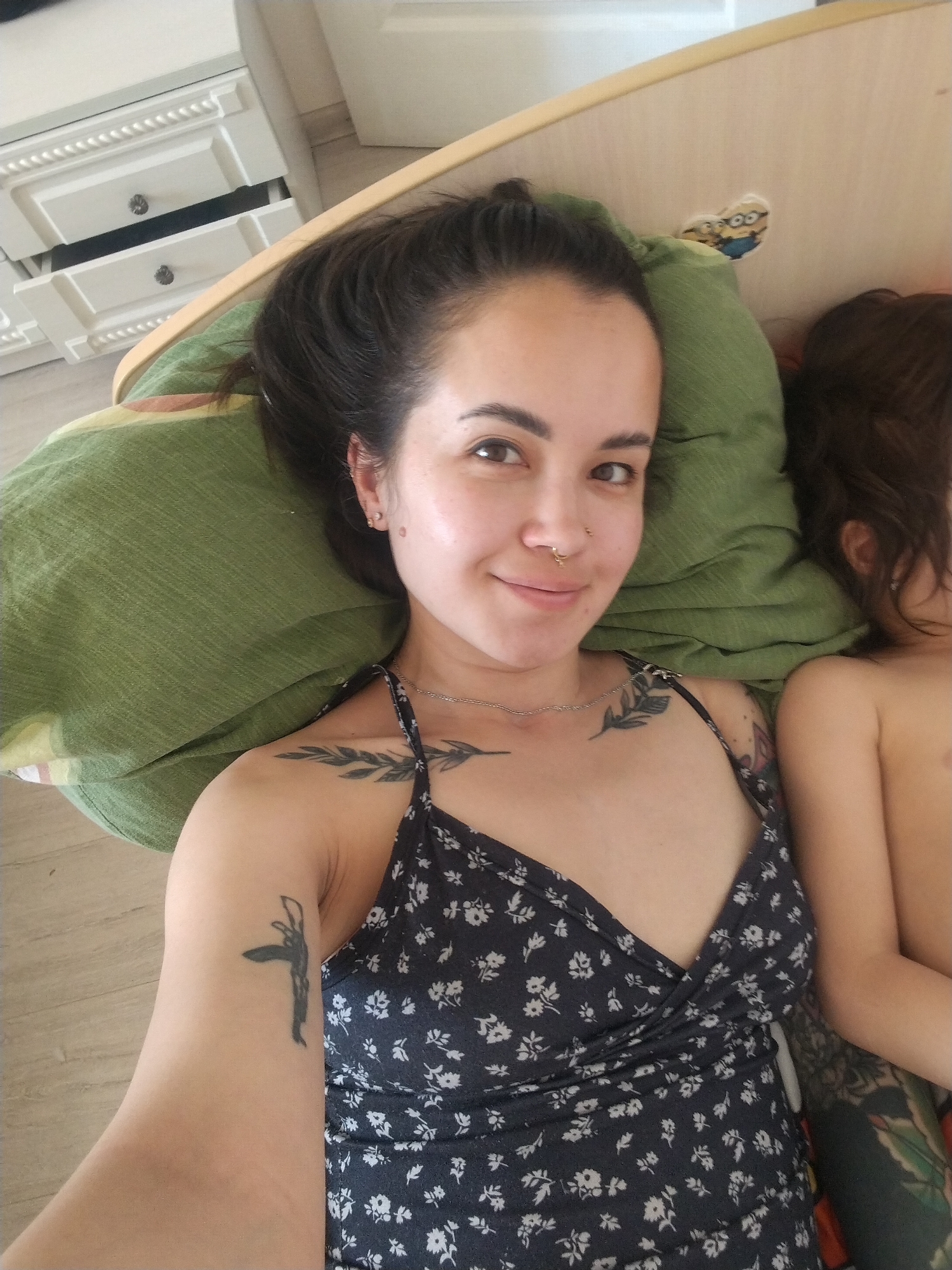 A selfie taken indoors shows a woman lying on a bed with a green pillow. She has dark hair pulled back, a nose ring, and tattoos on her upper arm, chest, and shoulder. She wears a dark floral print dress. A young child with brown hair lies beside her. A white dresser is visible in the background, and a sticker of cartoon characters is affixed to the headboard. The location and city of Noviy, Russia cannot be verified with the given image.
