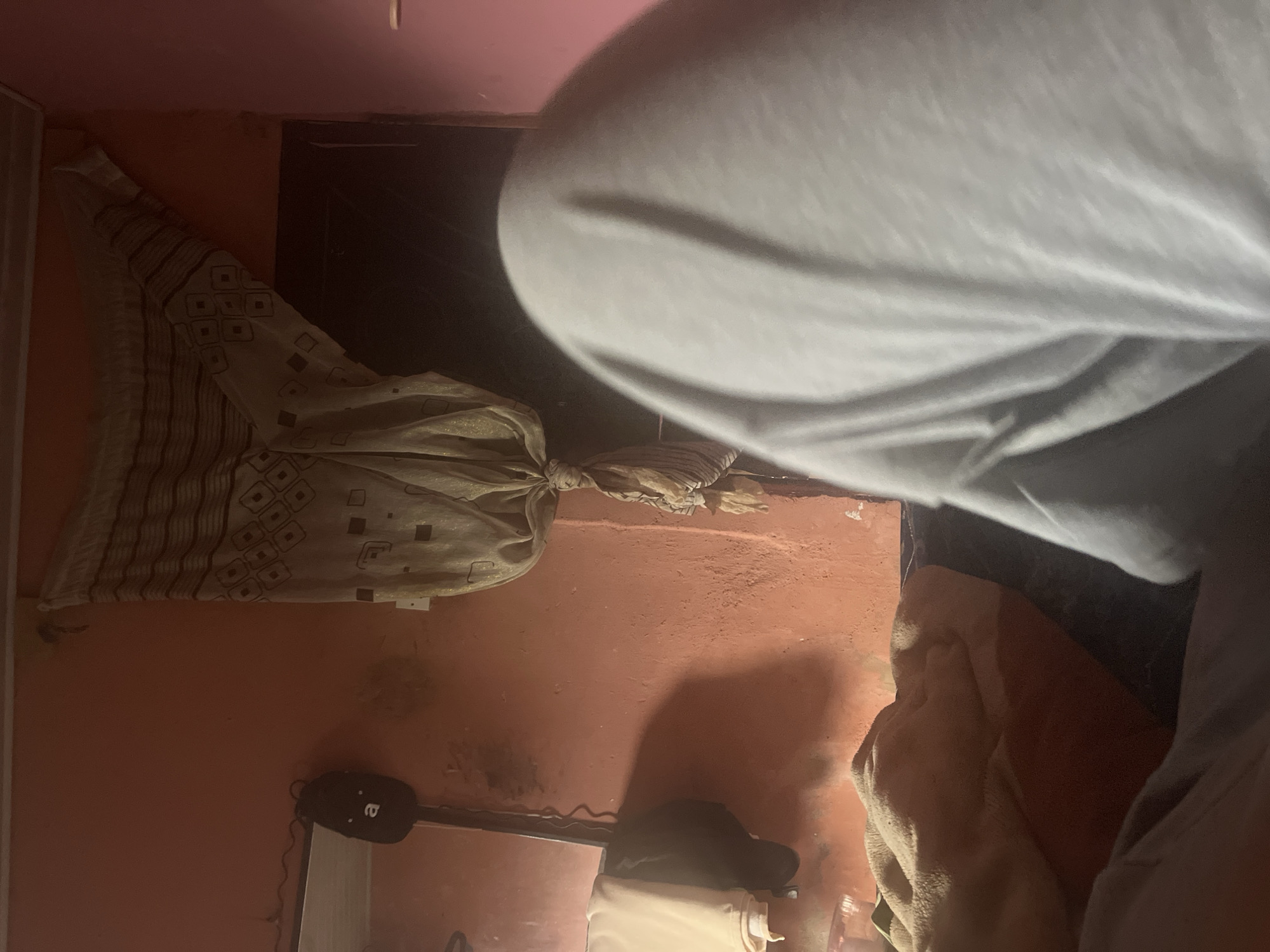 The image shows a close-up view of a person in a dimly lit room. The person is wearing a light-colored, possibly grey, hooded sweatshirt. Their head and upper body are prominent in the foreground, obscuring much of the background. A patterned fabric, possibly a scarf or garment with a diamond and stripe design, is tied in the middle and draped on the left side of the frame. The wall behind the fabric is a peachy or terracotta color, with some dark smudges or marks. On the lower part of the wall, a black object with a white circular emblem resembling a face or logo is visible, attached to some wires. Further down, a shelf or surface is present, with a white object on it and a darker object, possibly a cap, beside it. The lighting suggests it might be evening or night. The overall impression is of a personal, indoor space, possibly a bedroom or living area. There is no indication of weather or specific time of day beyond the artificial lighting. The emotional tone is neutral, focusing on the physical presence of the person and the surrounding objects.