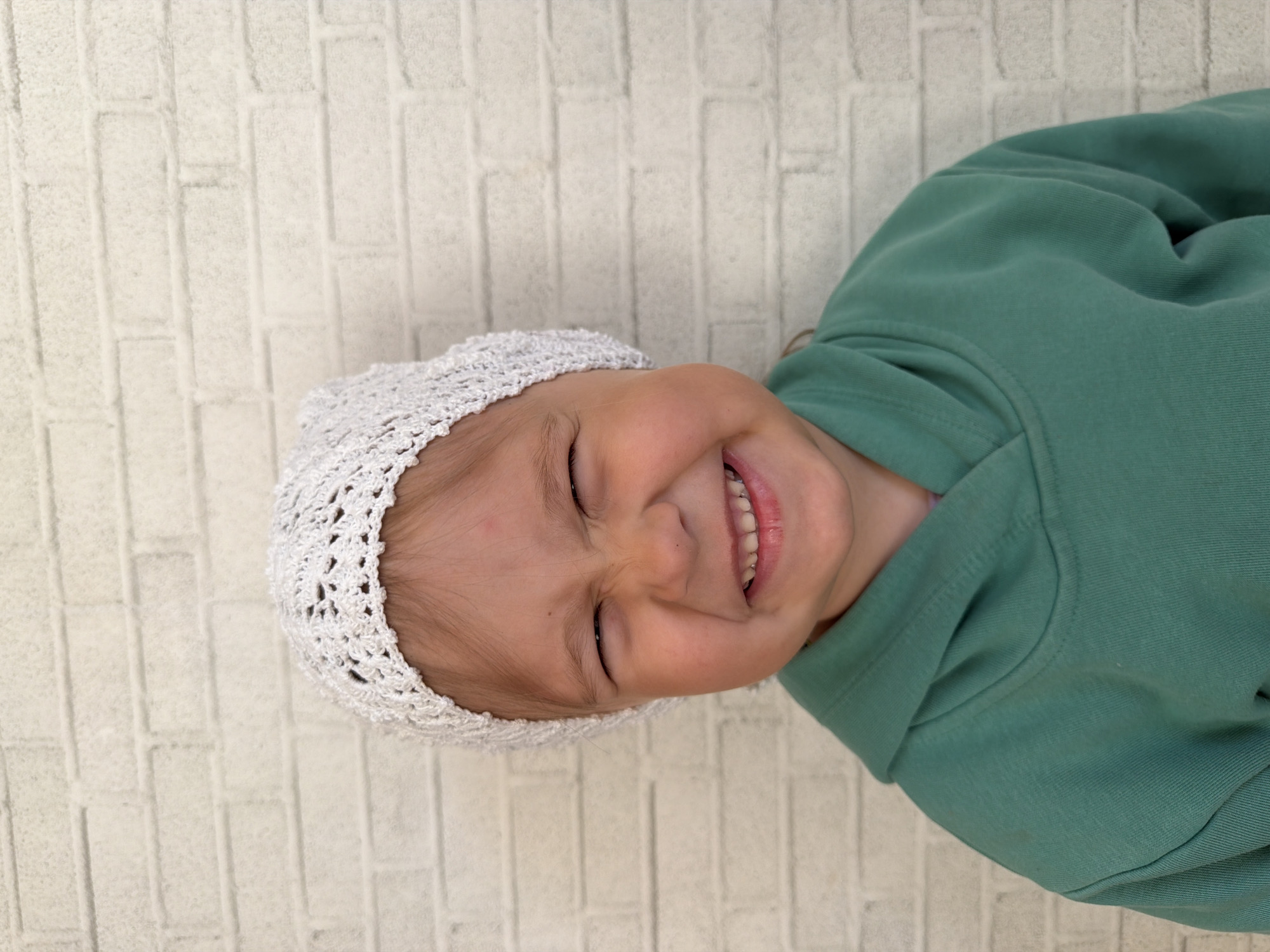 A young child wearing a white crocheted hat and a green hoodie is smiling with their eyes closed against a white brick wall. The location is likely Chaikovsky, Russia.