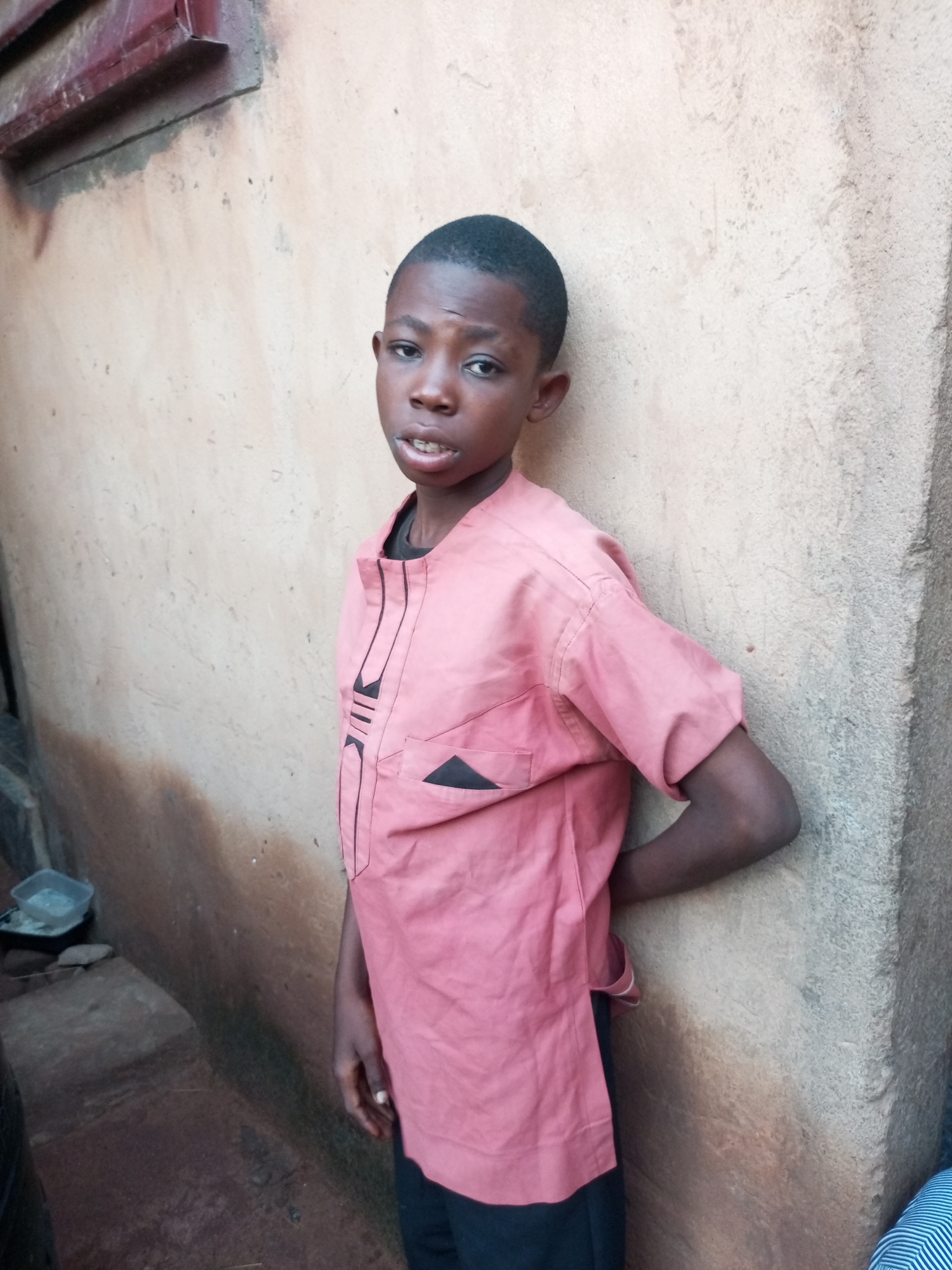 In Tempure, Nigeria, a young boy stands leaning against a textured, light-brown wall. He is wearing a salmon-colored traditional African tunic with black trim and accents, including a triangular patch on the left chest pocket and vertical stripes along the placket. His dark hair is neatly cut, and his mouth is slightly open. His left hand is behind his back, while his right arm is bent at the elbow. The wall appears weathered, with some dark staining and lighter patches. To the left of the boy, the ground is dark and uneven, with a small plastic container of what appears to be water or a liquid, and some rocks visible. Further to the left, a section of a building with a reddish-brown metal overhang is visible. The lighting suggests it is daytime.