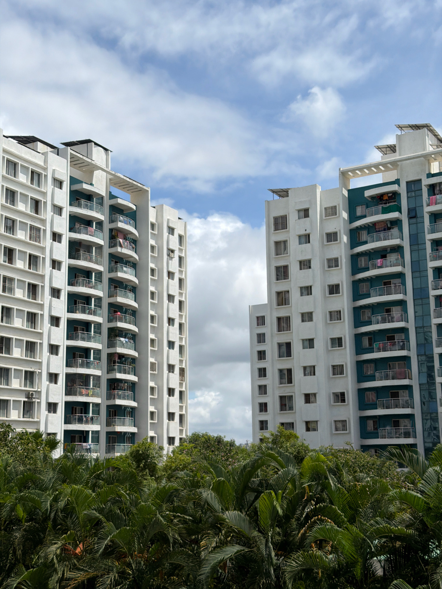 Two tall apartment buildings stand side-by-side, their white facades punctuated by rows of windows and balconies. The building on the left has a more uniform design with numerous square windows. The building on the right features balconies with curved railings and a teal-colored accent on some sections. Laundry is visible hanging on several balconies, suggesting it is a residential area. Solar panels are visible on the roofs of both buildings. In the foreground, lush green palm trees create a natural barrier, obscuring the lower portions of the buildings. The sky above is a mix of blue and white clouds, indicating fair weather. This scene is likely located in a modern urban area of Pune, India, given the style of the architecture and the presence of tropical vegetation.