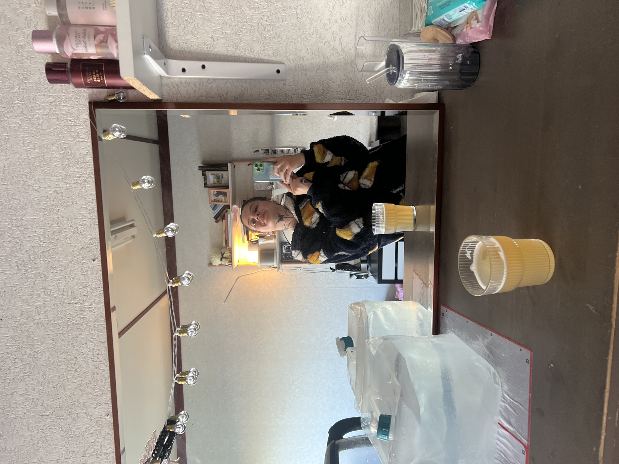 In Genemuiden, Netherlands, a woman takes a selfie in a mirror. She is wearing a dark, fuzzy bathrobe with a pattern of yellow and white shapes resembling mugs of beer. Her reflection shows her holding a smartphone and facing the mirror. There are lightbulbs strung along the top of the mirror. On a shelf to the right of the mirror, there are several bottles of perfume. In the foreground, on a dark wooden counter, there are two large, clear bags of water with spigots. Next to these, there is a glass filled with a light-colored liquid, likely beer. To the right of the glass, there is a grey glass tumbler with a straw. The background appears to be a wall with a textured surface.