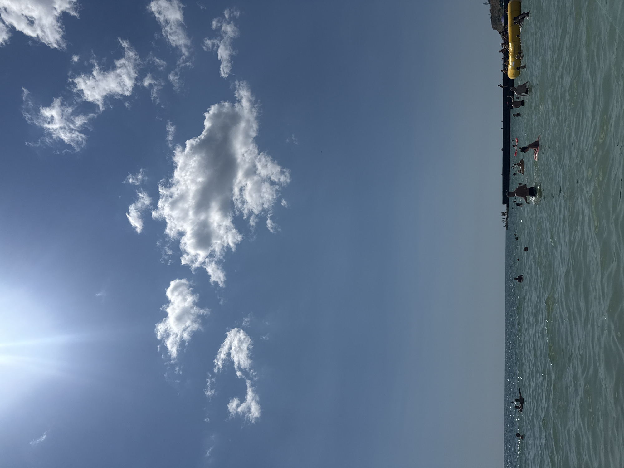 A bright, sunny day at a crowded beach in Odesa, Ukraine, is captured from a low angle in the water. The sky is a vibrant blue, with scattered white clouds, and the sun shines intensely from the upper left, creating lens flare. The sea is a clear, shimmering turquoise, with gentle waves.
In the foreground, numerous people are enjoying the water. Some are swimming, while others are floating on inflatable toys, including a prominent yellow inflatable structure on the right side of the image. The people are of various ages and genders, appearing relaxed and engaged in recreational activities.
In the background, a long, dark breakwater extends into the sea, with more people gathered on it and on the shore beyond. Further in the distance, a hillside with some structures is visible. The overall impression is one of a lively and popular summer beach scene.