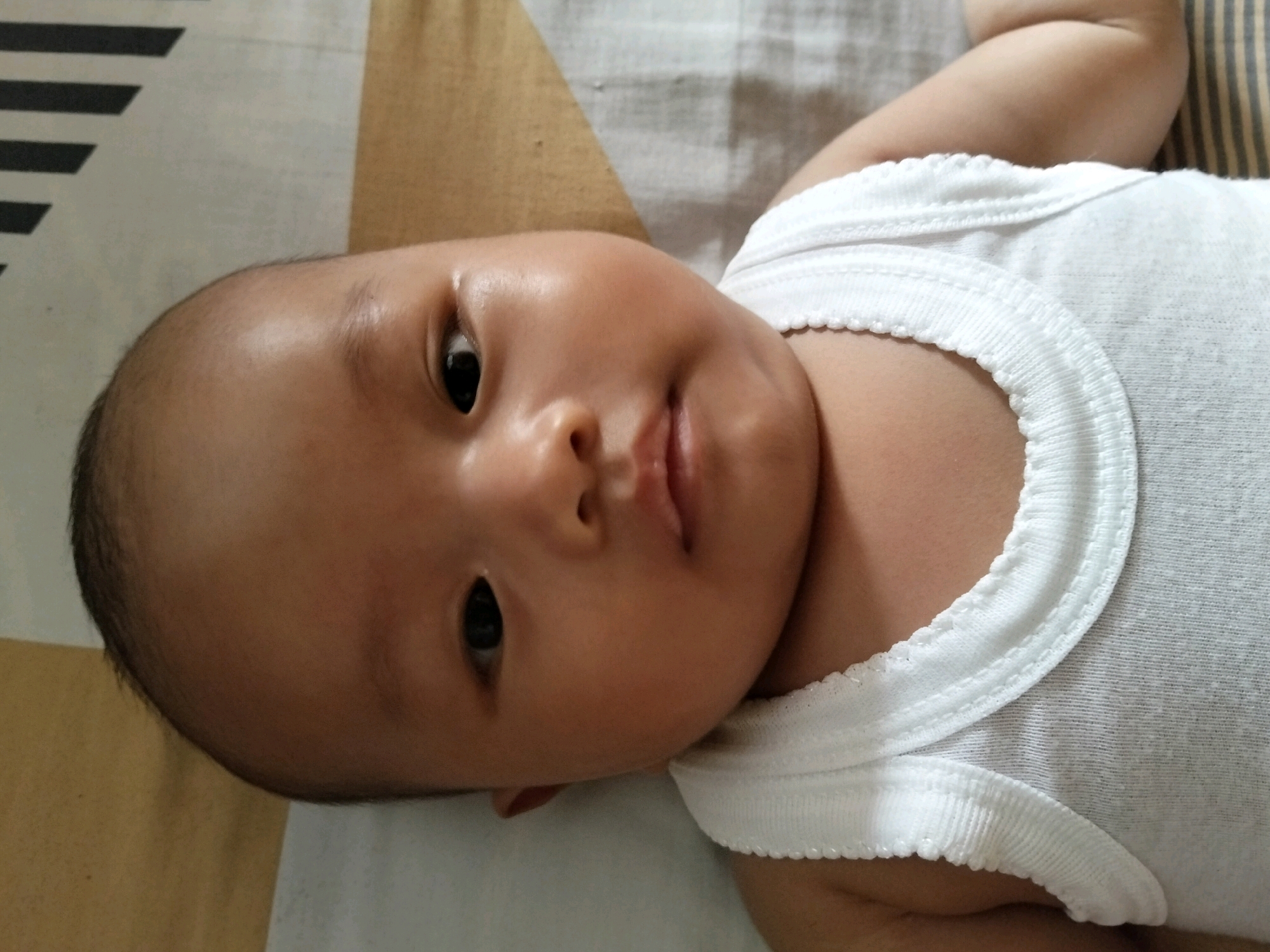 A close-up, low-angle shot captures a baby lying on a patterned surface. The baby is wearing a white sleeveless onesie with a scalloped edge. The baby's head is turned to the left, with their face prominently featured. Their skin is smooth and their eyes are open. The background consists of a light-colored textile with a diagonal pattern of dark grey and black stripes on the left side and a tan-colored area on the right. The lighting suggests an indoor setting. The image appears to be taken at a residence in Gempolan, Indonesia.