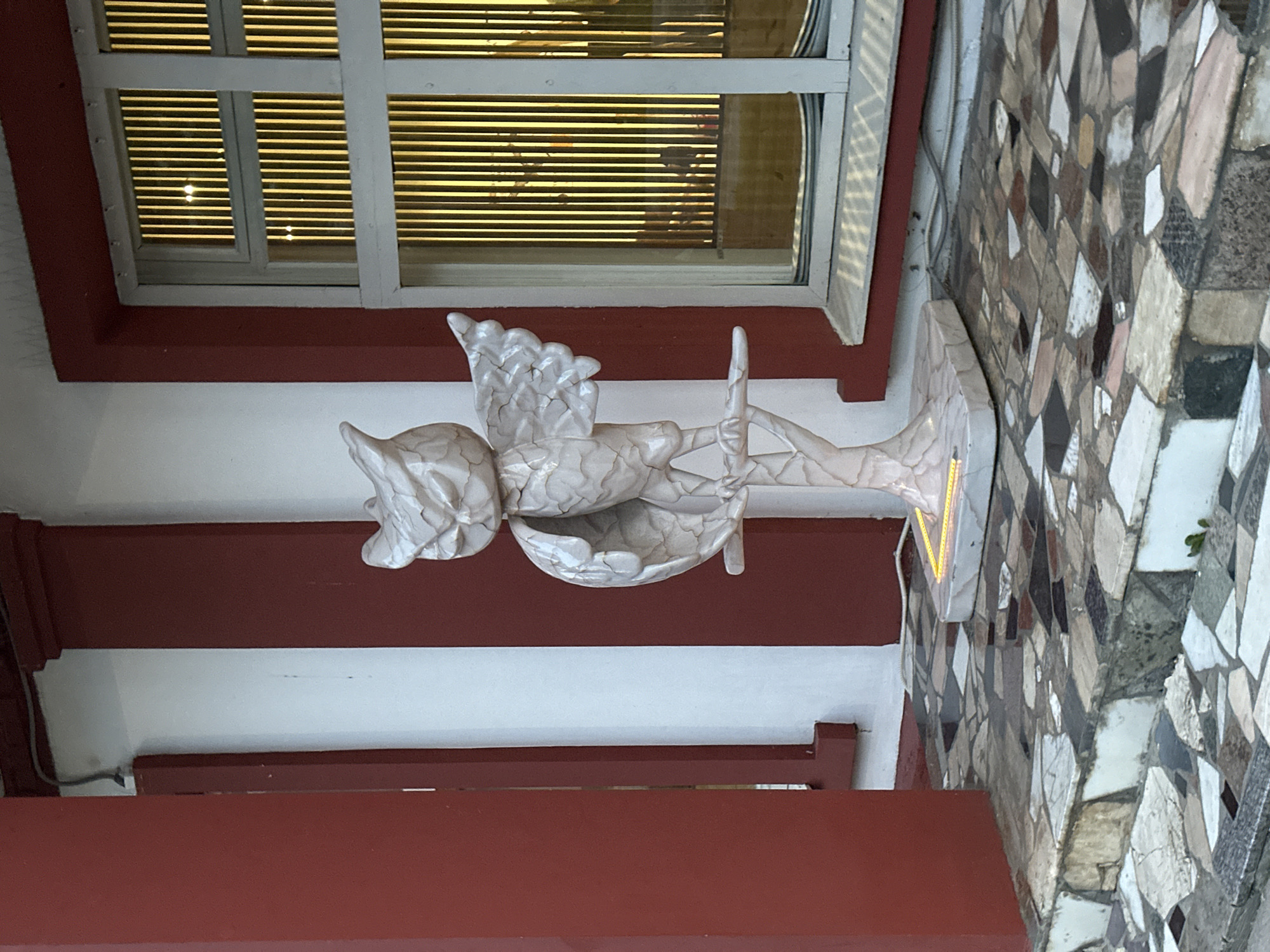 The image, taken in Moscow, Russia, captures a static outdoor scene featuring architectural elements and a decorative statue. In the center, a white, cracked ceramic or plaster statue of a winged figure, possibly an angel or cherub, holds a bowl-like vessel. The statue rests on a pedestal, positioned against a white wall with a prominent red-brown painted base and trim.
Above the statue, a window with white frames is visible, equipped with venetian blinds that are partially open, allowing horizontal streaks of yellow light to filter through, suggesting an interior light source or reflection. To the right of the statue, a wall is covered in an intricate mosaic pattern. This mosaic is composed of numerous irregularly shaped tiles in various earthy tones, including brown, white, grey, black, and beige.
The lighting indicates it is daytime. No specific activity is taking place, and there is no visible text. The scene conveys a sense of ornate detail and aged charm within a building's exterior or courtyard setting.