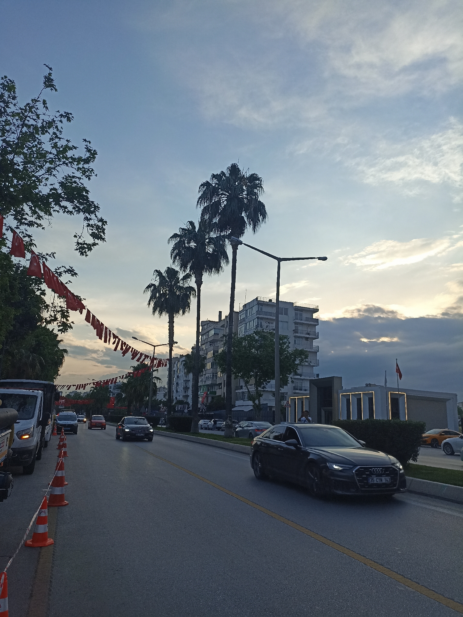 The image depicts a street scene in Yenişehir, Turkey, at dusk. The roadway is lined with palm trees and buildings, with red Turkish flags strung across the street. Cars are traveling on the road. Orange traffic cones are visible on the left side of the road. The sky is a mix of blue and cloud formations.
