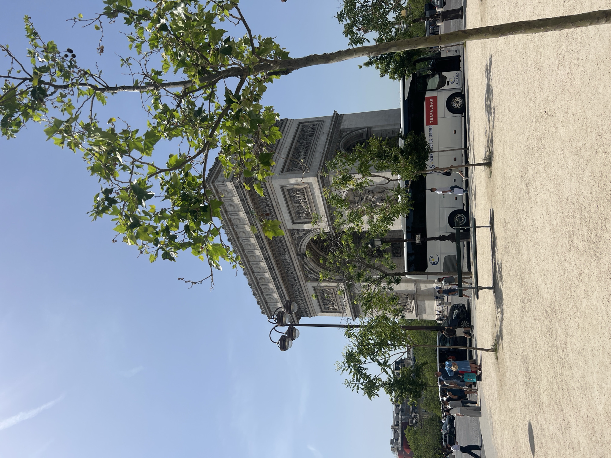 The image shows the Arc de Triomphe in Paris, France, with a Trafalgar tour bus parked alongside it. People are present, walking and standing near the bus and the monument. The sky is clear blue, and trees frame the view of the Arc. A lamppost stands on the left side of the image. The ground is covered with light-colored gravel. Buildings are visible in the background, indicating an urban environment.