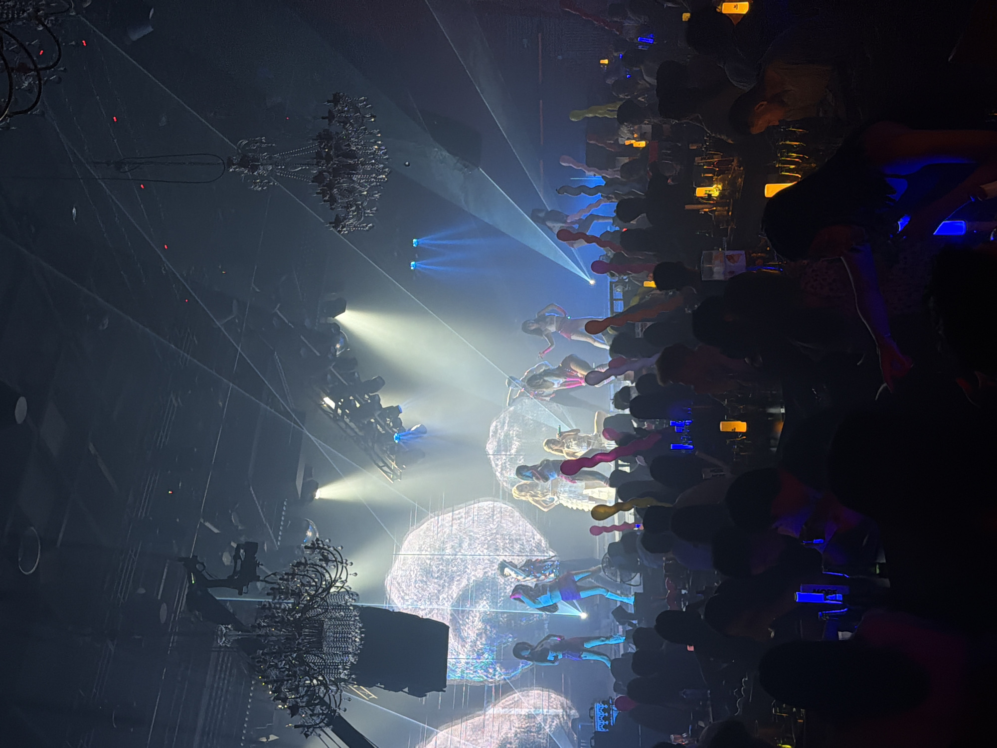 The image depicts an indoor performance venue, likely a club or concert hall, in Jiangbeicheng Street, China. The scene is dimly lit, with a stage area illuminated by bright spotlights and theatrical lighting. On stage, a group of performers, predominantly women dressed in revealing costumes, are engaged in a performance. They are positioned in various poses, suggesting dancing or a staged presentation. Behind them, large circular projections, possibly screens or illuminated artwork, serve as a backdrop.
In the foreground, a seated audience occupies tables, their faces obscured by the low light and shadows. The audience members are holding various objects, including illuminated sticks or glow sticks, indicating an active and engaged crowd. The environment is characterized by a dark, smoky atmosphere, with chandeliers suspended from the ceiling adding a touch of opulence. The overall impression is one of a lively entertainment event with a focus on performance and audience interaction.