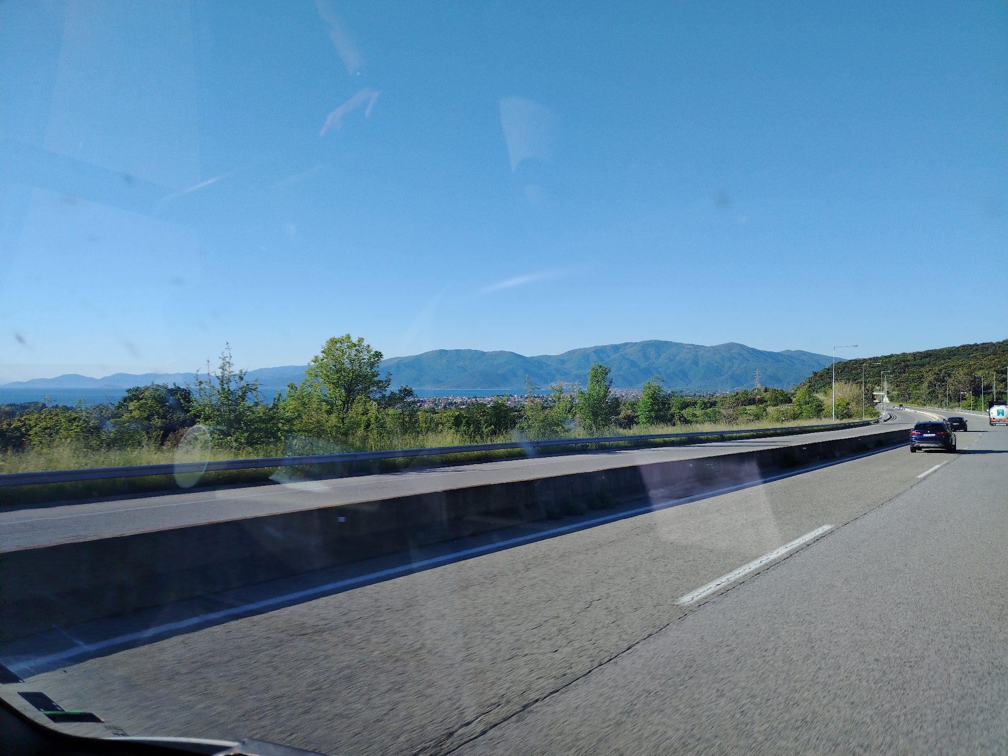 The image depicts a view from a vehicle traveling on a highway in Ασπροβάλτα, Greece. The sky is clear and blue. Mountains are visible in the distance. A concrete barrier separates the two lanes of the highway. A few cars are visible on the right side of the road. The foliage on the side of the road includes trees and green plants. A section of the town is also seen between the mountains and the greenery. There's also a possible reflection visible at the top of the image due to the shot being captured through the vehicle's window.
