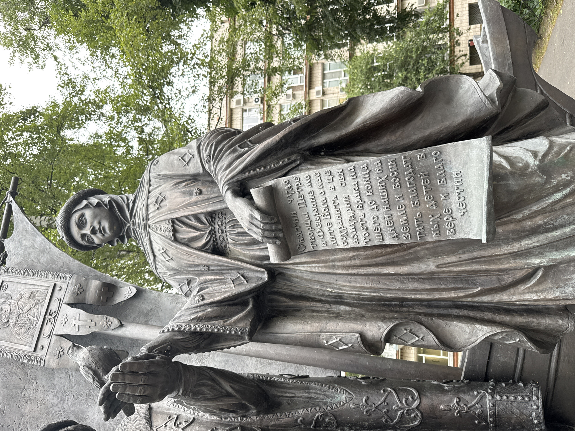 The image shows a bronze statue of a woman in traditional clothing holding a scroll. The statue is located in the city of Ladozhskoe Ozero, Russia, possibly as part of a larger monument or memorial. The woman is depicted with a serene expression and is wearing a head covering and a flowing robe adorned with decorative patterns. The scroll she holds contains inscribed text in an unfamiliar script, suggesting it may be of historical or religious significance. The statue is detailed, with realistic textures and folds in the fabric of her clothing. Behind the statue, a building with multiple windows and air conditioning units is visible, surrounded by lush green trees. The overall setting appears to be an outdoor public space, likely a park or square, given the presence of trees and paved ground. The lighting suggests it is daytime, with overcast skies.