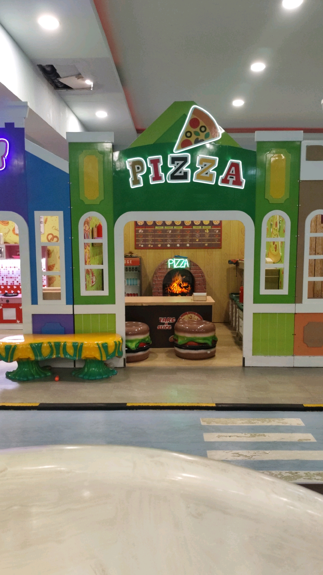 This is a medium shot, eye-level, interior photograph of a colorful, child-friendly pizza play area. The play area is designed to resemble a miniature town with buildings of various bright colors, including blue, green, and yellow. A prominent sign above the entrance to the pizza shop reads "PIZZA" in large, illuminated neon letters, accompanied by a graphic of a pizza slice.
The pizza shop itself features a faux brick oven with a realistic-looking fire, a counter with a menu board displaying various pizza options, and a refrigerator unit. Two large, inflatable hamburger-shaped chairs are placed in front of the counter. To the left of the pizza shop, there is a play kitchen area with visible appliances and decorations like pretzels and pastries. In the foreground, a table with a yellow tablecloth and green, decorative legs is set up. The floor is a light-colored tiled surface with blue painted accents and what appears to be a stylized crosswalk. The ceiling above shows evidence of some damage, with a section of the ceiling tile missing, revealing darkness behind it. The lighting appears to be from overhead fluorescent fixtures, casting a bright, even illumination on the scene. There are no people visible in the photograph. The overall impression is of a commercial entertainment space designed for young children.
This play area is located within the Trans Studio Mall in Bandung City, Indonesia.