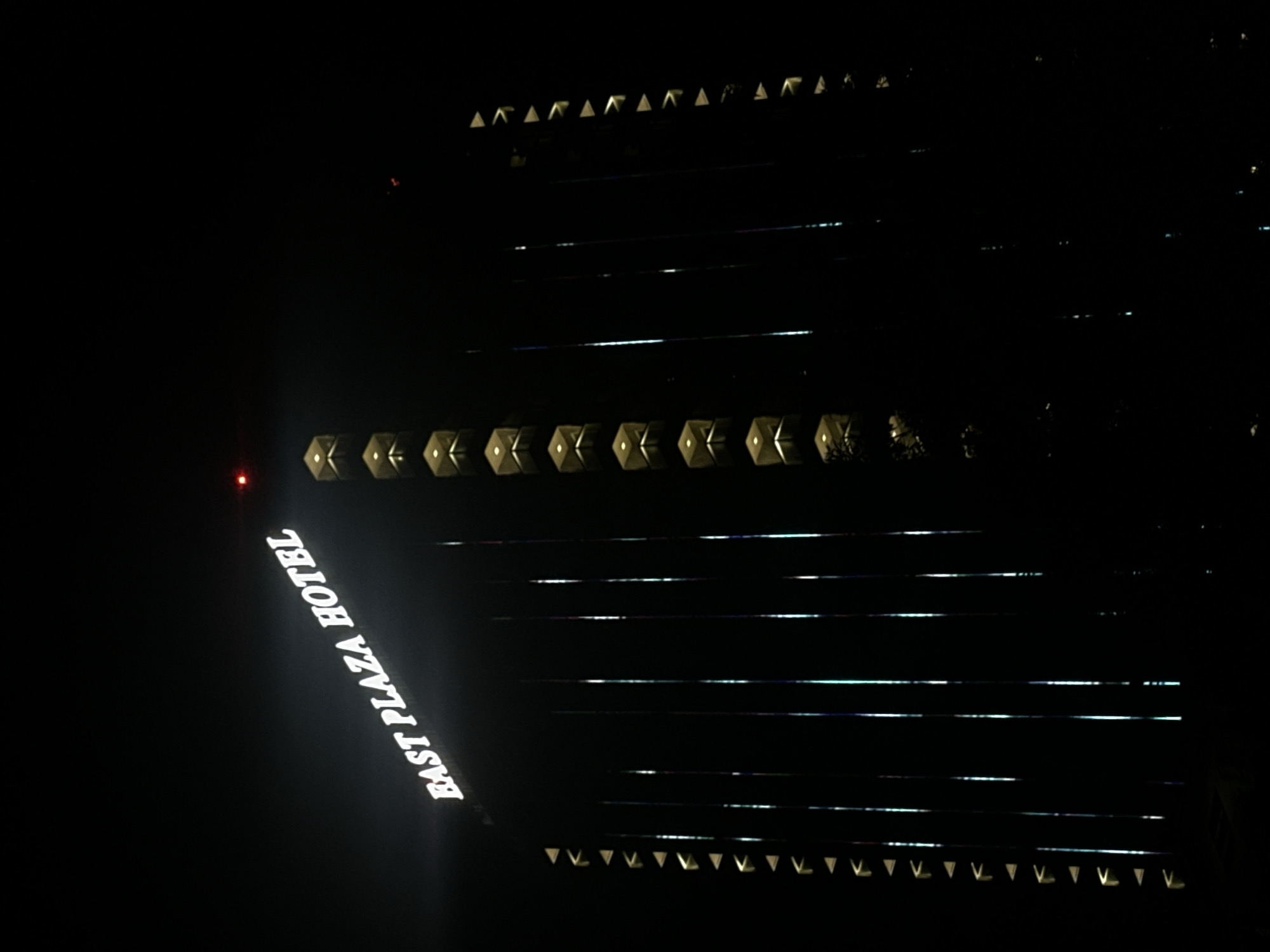 The image shows the East Plaza Hotel at night in Sofia, Bulgaria. The hotel is a tall, dark building with illuminated signs and lights visible on its exterior. The "EAST PLAZA HOTEL" sign is prominently displayed in white, illuminated lettering near the top of the building. Rows of triangular-shaped lights run down the sides of the building, creating a geometric pattern against the dark facade. Vertical lines of light, possibly from illuminated windows or architectural features, are also visible on the building's surface. A small red light is visible at the very top of the building, potentially a navigational warning light. The overall scene is dark, with the building being the primary source of light. The surrounding environment is obscured by darkness.