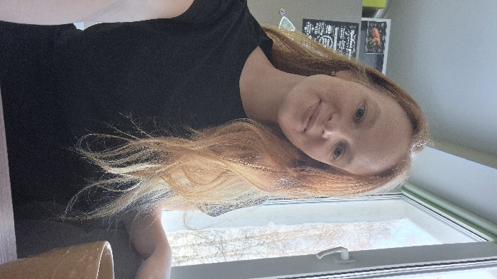 The scene is an interior view within the city of Chaikovsky, Russia. A young adult female with long, wavy, reddish-blonde hair and light-colored eyes is positioned centrally, gazing directly at the viewer with a slight smile. She is wearing a black short-sleeved t-shirt. To her left, a light-colored refrigerator is partially visible, adorned with a dark magnetic board displaying white text and a smaller magnet depicting a roasted fowl. To her right, a white-framed window with a visible white handle reveals bare tree branches against a bright sky in the exterior. A tan ceramic mug is partially visible on a dark horizontal surface in the lower right foreground, where the subject's right arm is resting.