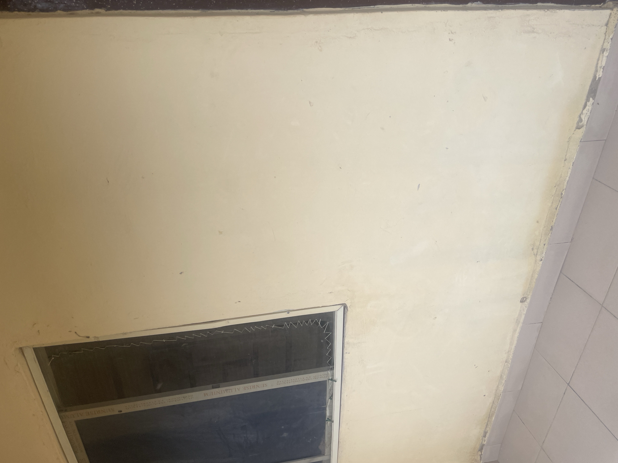 The image shows a close-up view of a cream-colored wall with a vent or window at the bottom left. The wall appears to be an interior wall, possibly in a residential or commercial building. The vent has a metal grate and what looks like a dark screen behind it. There is some staining or discoloration on the wall, particularly along the right edge where it meets a tiled surface. The lighting suggests it is daytime, but no specific time can be determined. The scene is static, with no people or activity visible. The overall impression is one of a somewhat dated or worn interior. The text "SUNRISE ALUMINUM" is visible on a strip within the vent. No other text is legible. The location context is Jalingo, Nigeria, but this is not visually evident from the image itself.