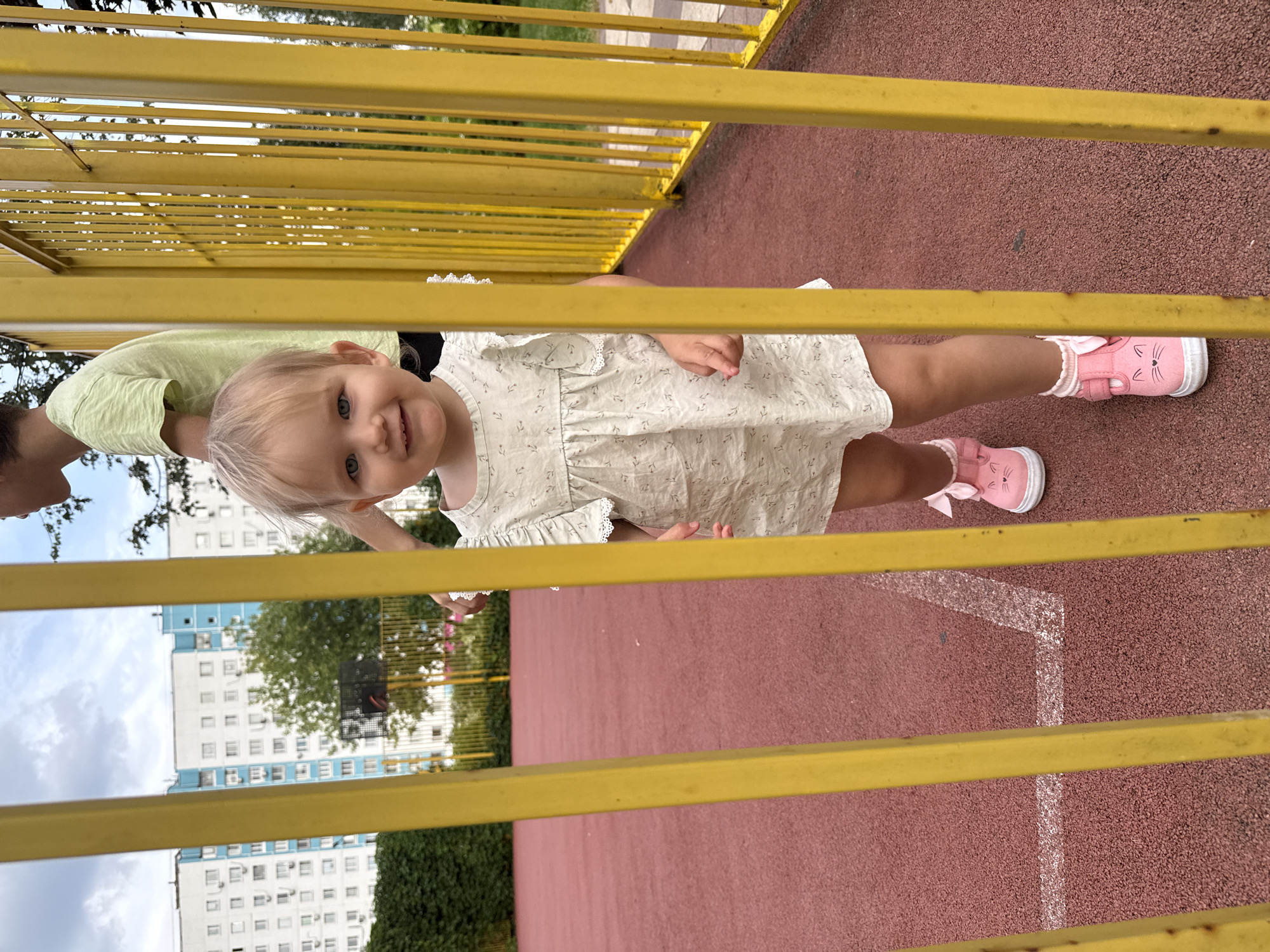 In Moscow, Russia, a young girl with blonde hair and blue eyes, wearing a light-colored dress with a floral pattern and pink cat-themed shoes, stands behind a yellow metal fence. She is smiling and looking at the camera. Behind her, a person in a green t-shirt is partially visible. The background shows a residential building with blue and white accents, trees, and a basketball hoop. The ground is a red, textured surface, possibly a sports court, with a white line marking. The sky is cloudy.