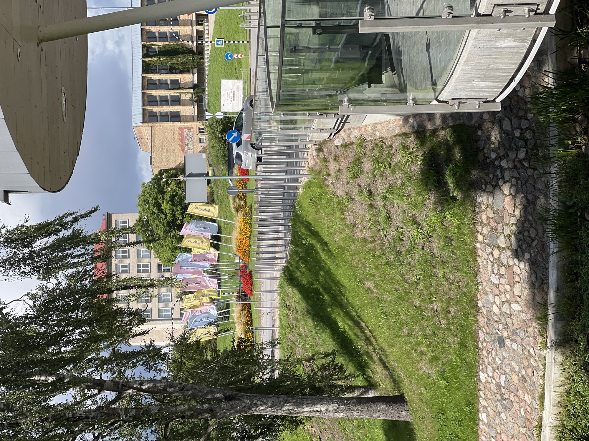 The image captures a view of the Latvian Academy of Culture's building in Saldus, Latvia. The foreground features a modern, curved glass and concrete structure, likely an entrance or viewing area. To the left, a grassy slope with a stone retaining wall leads up towards a street. A prominent birch tree with sparse leaves stands on the left side of the frame, casting shadows on the grass.
In the middle ground, a circular arrangement of colorful flags in shades of yellow, pink, and blue are displayed on poles. These flags appear to be decorative and are set against a backdrop of vibrant red, yellow, and orange flowers. Behind the flags, a modern, light-colored building with numerous windows, characteristic of institutional architecture, is visible. Further in the background, another brick building with a blue roof and a facade covered in ivy is present. A silver car is seen driving on the road in front of the buildings.
Several traffic signs, including directional arrows and pedestrian crossing signs, are positioned along the roadside, indicating a regulated area. The overall scene suggests a public space, possibly a campus or cultural center, with elements of modern architecture, landscaping, and outdoor decorations. The sky is overcast with some patches of brighter sky visible.