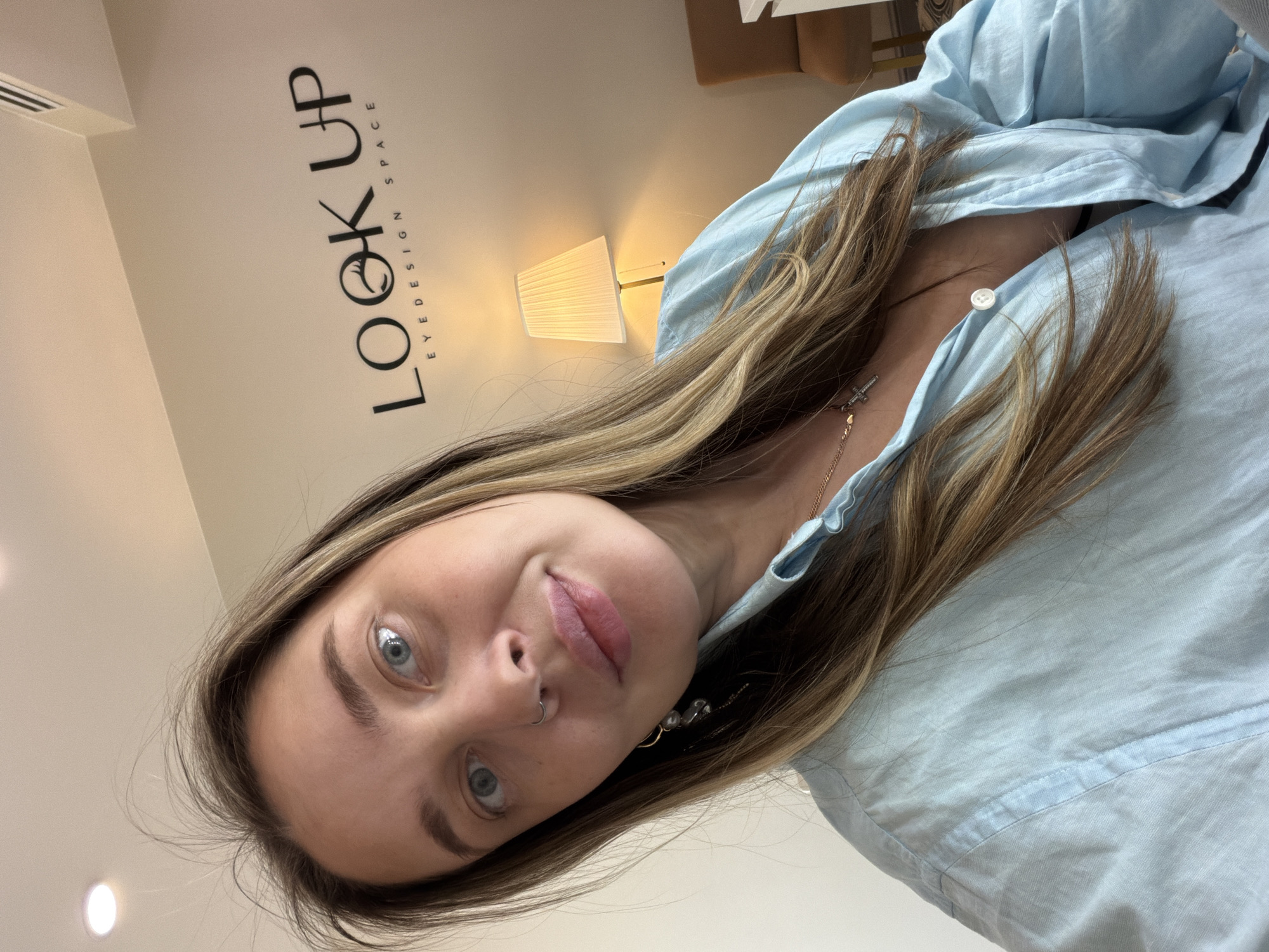 A young woman with blonde and brown hair, blue eyes, and a nose piercing is posing in front of a wall that says "LOOK UP EYEDESIGN SPACE" in black lettering. The image was likely taken inside an interior space that provides eyedesign services, as indicated by the logo. A lamp can be seen in the background. The woman is wearing a light blue collared shirt and a necklace with a cross pendant. There is no evidence that the photo was taken in Moscow, Russia. The location is an eyedesign space.