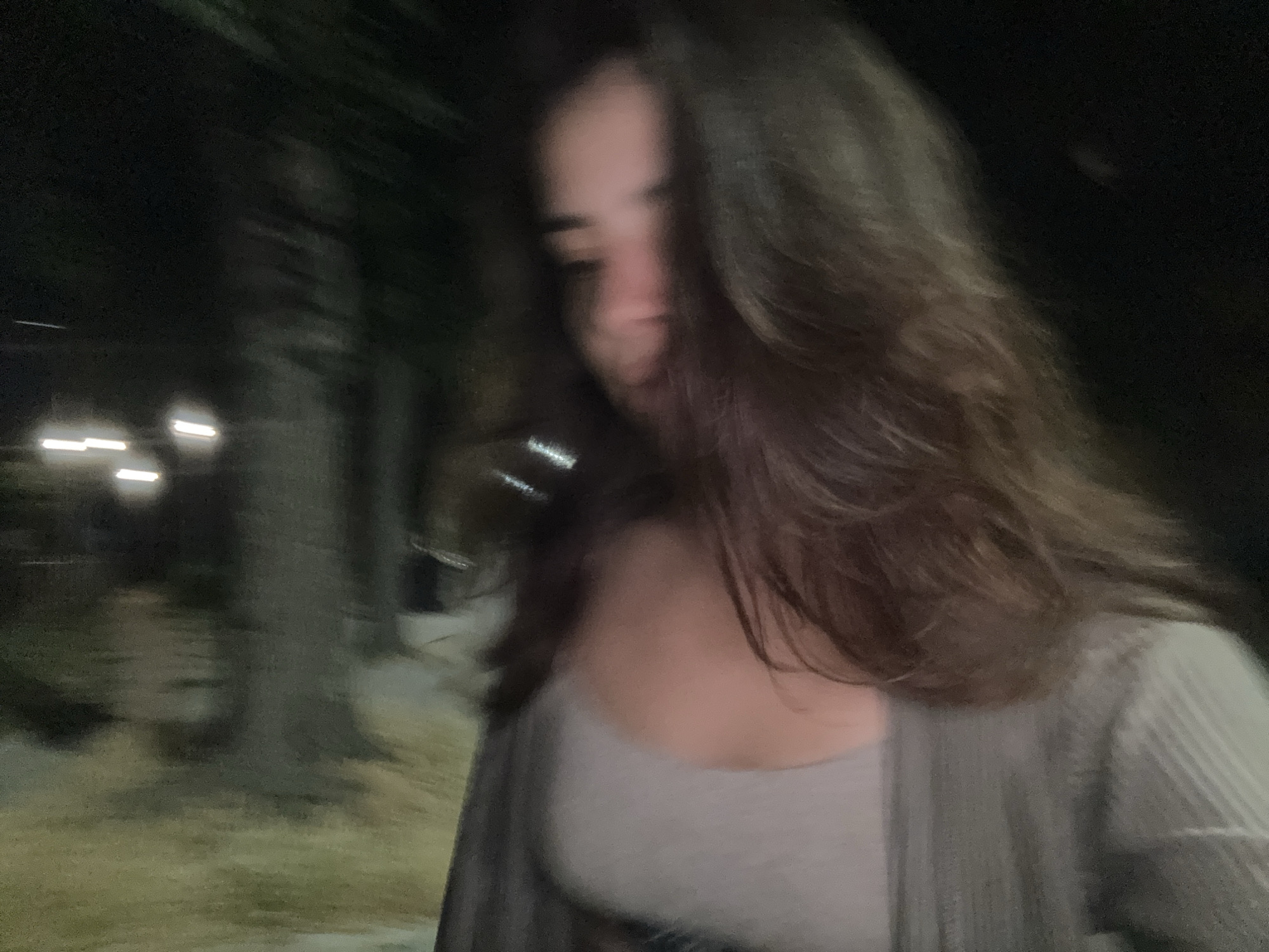 The media file shows a person with long, dark hair looking down and to the side. They are wearing a light-colored top and a grey cardigan. The background is dark and blurry, with some faint lights visible, suggesting it is nighttime. Trees are also visible in the background, giving a sense of an outdoor setting, possibly a park or street with trees. The overall image quality is low, with motion blur and low light conditions. The subject's face is partially obscured and out of focus. There are no visible text elements in the image. The setting appears to be Plovdiv, Bulgaria, but there are no specific landmarks or cues to confirm this definitively from the image itself. The activity taking place is not clear due to the blurriness and the subject's downcast gaze. The emotion conveyed is ambiguous due to the lack of clear facial features.