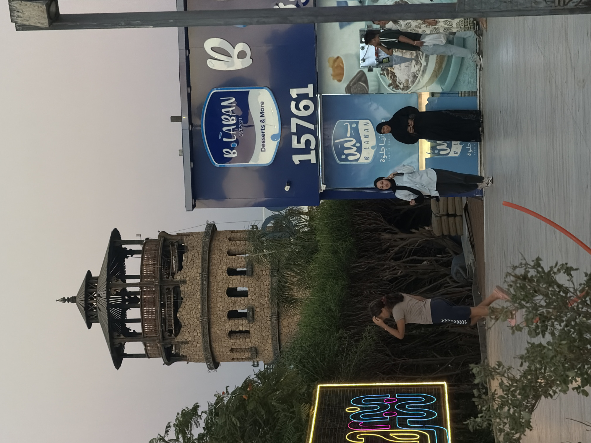 In Sharm El Sheikh, Egypt, a cobblestone tower with a wooden upper structure stands against a pale sky. To its right, a blue building features the logo "B.LABAN EST. 2021 Desserts & More" and the number "15761". Two women in dark headscarves and long dresses stand in front of the blue building, and a man in a green striped shirt and a woman in a patterned dress are also visible. A young woman with dark hair pulled back walks away from the camera towards the left side of the frame. In the foreground, a neon sign with swirling blue and pink shapes is illuminated. The ground is paved with large, light-colored tiles.