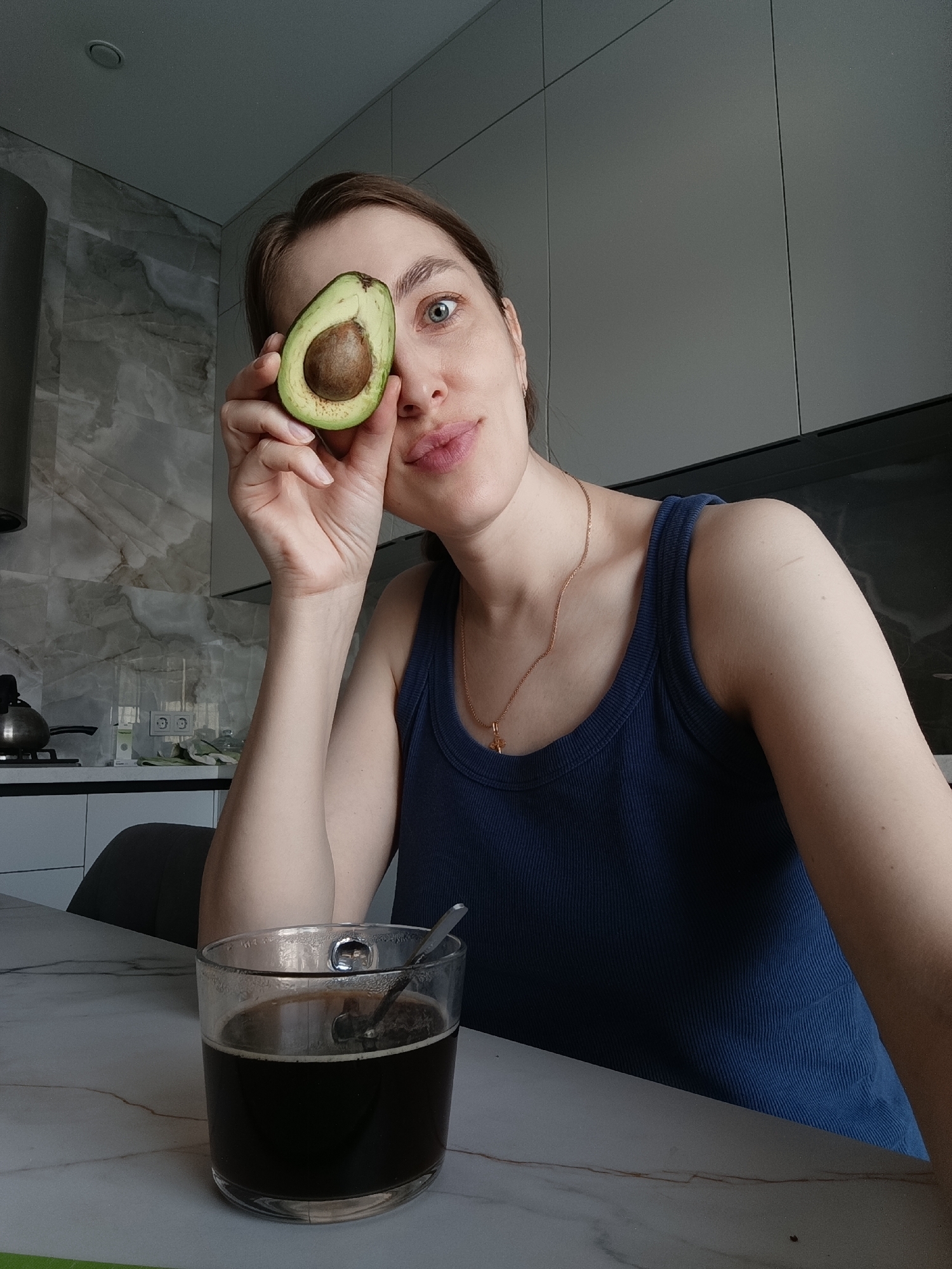 A woman sits at a table in a kitchen. She holds half an avocado up to her right eye. She is wearing a dark blue tank top and a gold necklace. A clear glass filled with dark liquid and a spoon sits in front of her on the table. In the background, there are kitchen cabinets and a stainless steel vent hood. The location is the city of Chaikovsky, Russia.