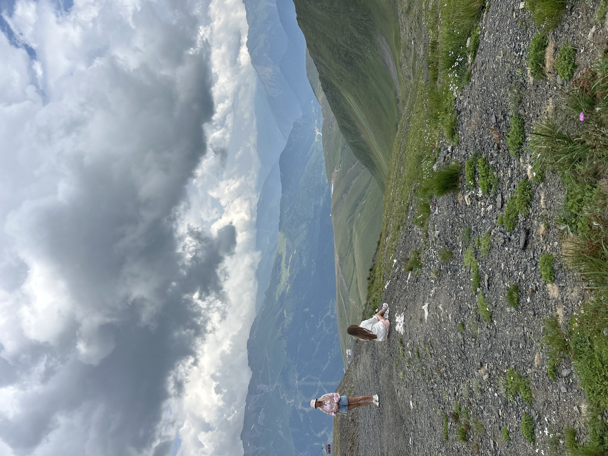 Two young females are positioned on a rocky slope overlooking a vast mountainous landscape. The female on the left, standing, wears a pink patterned jacket, light blue shorts, and a pink bucket hat. The female on the right is seated facing away from the camera, wearing a white top and pink shorts. The foreground consists of a dirt and gravel path with sparse green vegetation. The middle ground displays rolling green hills, and the background features layers of distant, hazy mountains under a dramatic, cloudy sky. A ski lift structure and cable are visible to the far left. The overall scene depicts a scenic mountain environment, likely in a resort area such as Lunnaya Polyana, Russia, given the presence of ski infrastructure.