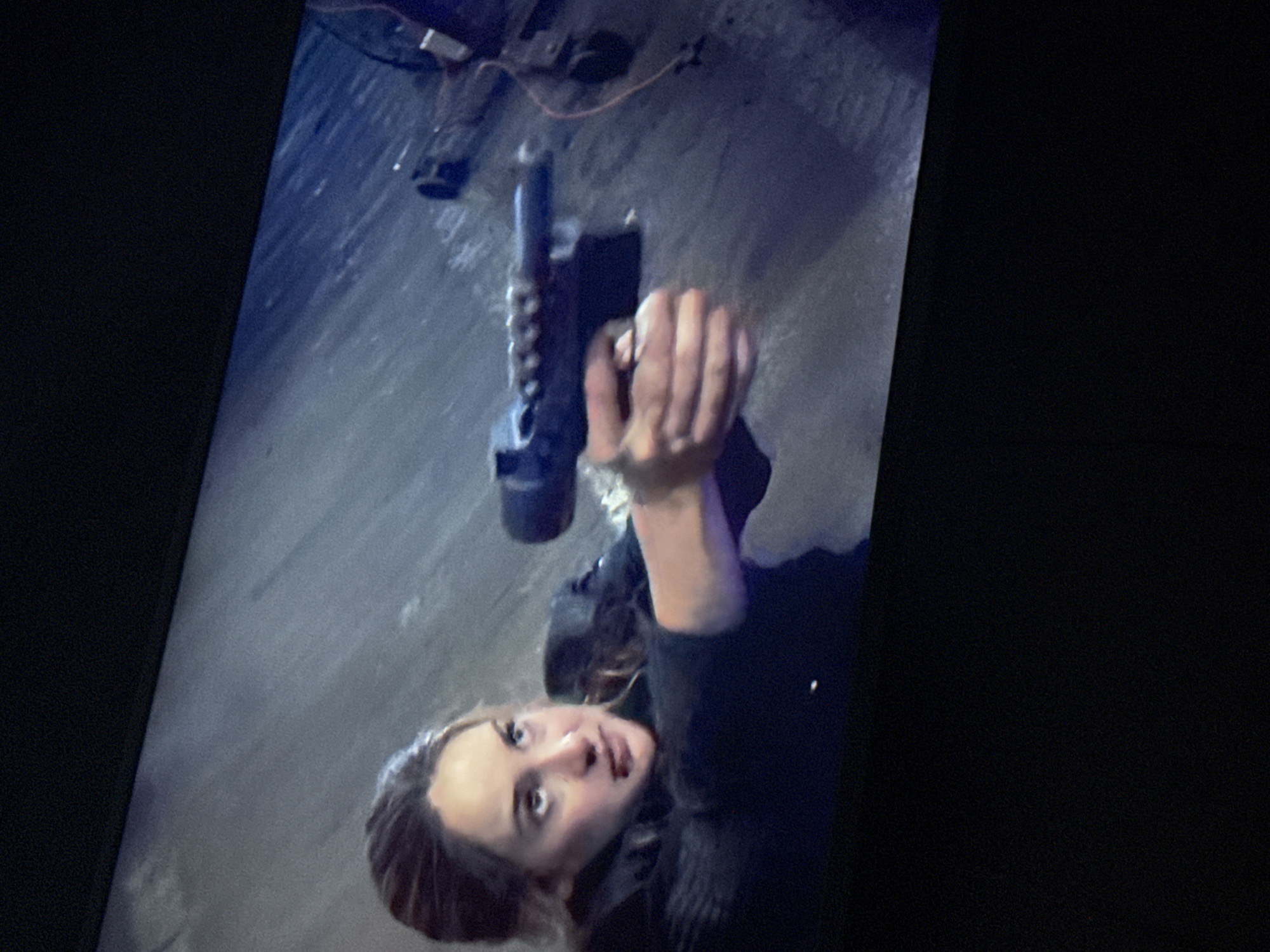 A woman is holding a gun upwards. The background is a pavement, with what appears to be a streetlamp and some cables on the ground. It is difficult to determine the location and interactions definitively.