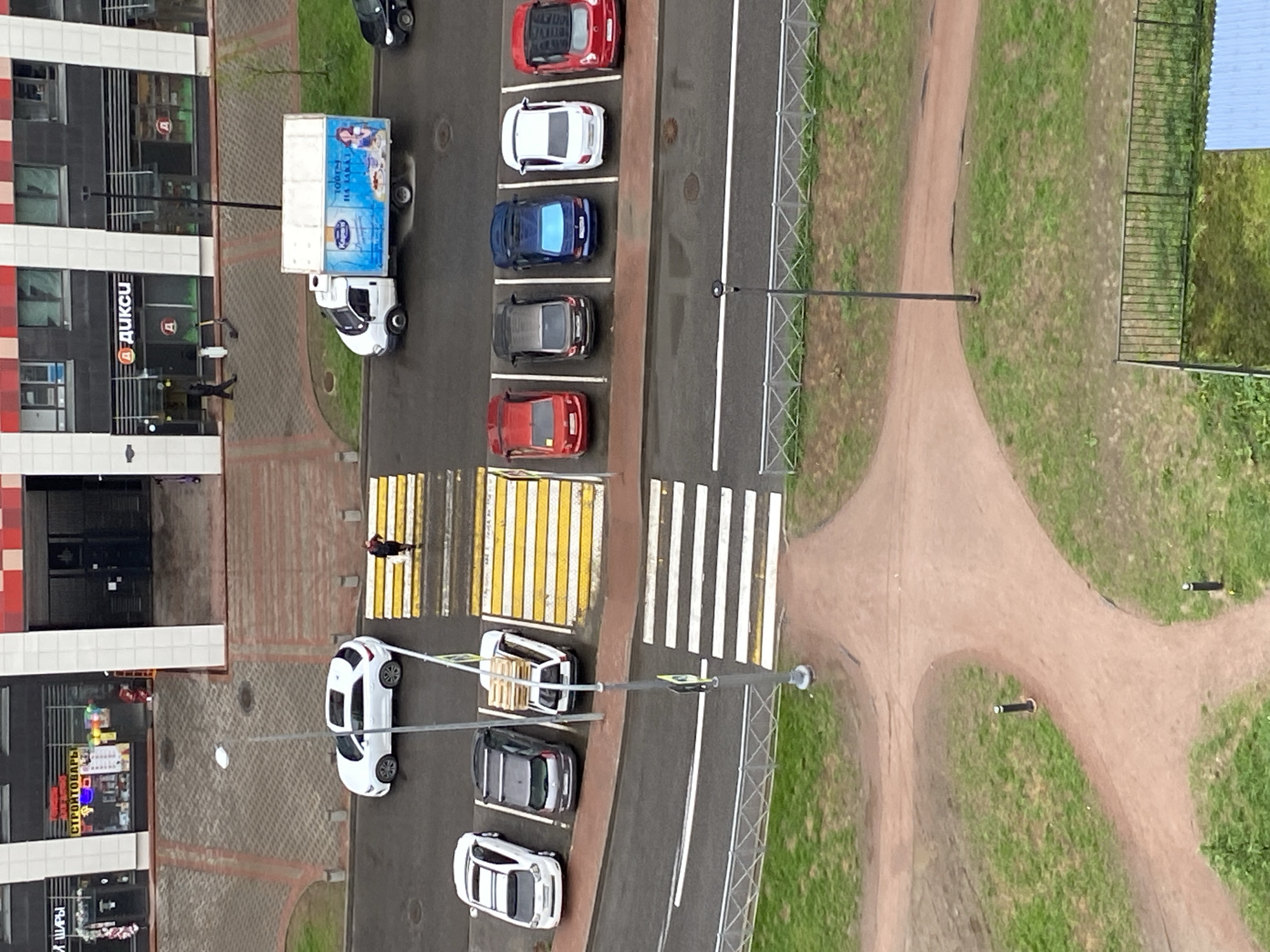 A high-angle, daytime photograph depicts a street scene in an urban setting, possibly in Russia. A building with commercial establishments, including a store identified as "ДИКСИ" (DIKSI), lines one side of the street. Several parked cars are visible along the road, including red, white, blue, and dark-colored vehicles. A delivery truck is parked near the building. Pedestrian crossings are marked on the street. A person is walking across one of the crosswalks. To the right of the road, a dirt path winds through a grassy area, with poles or posts along the path's edge. The scene suggests a blend of commercial and residential areas, with pedestrian traffic and vehicular parking.