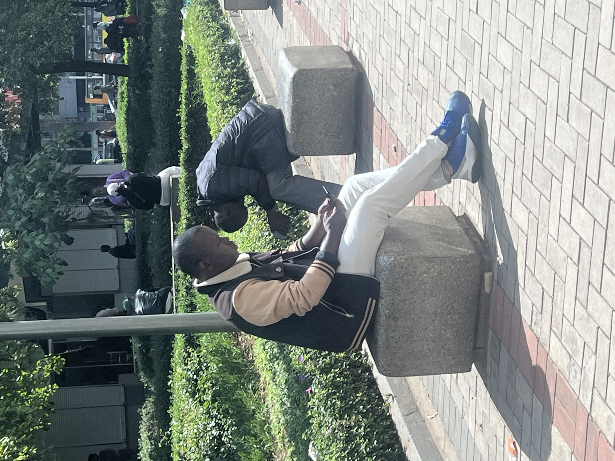 The image shows two young men in an outdoor setting, likely in Nairobi, Kenya. One man is seated on a concrete block with his legs outstretched, wearing white pants, blue and white sneakers, and a jacket with a tan and black color scheme. He is looking at something in his hands, possibly a phone. The second man is crouched down, also appearing to be looking at something in his hands. He's wearing a dark jacket. They appear to be situated next to a paved walkway. The background includes green bushes and trees, suggesting a park or urban green space. In the distance, other people are visible.