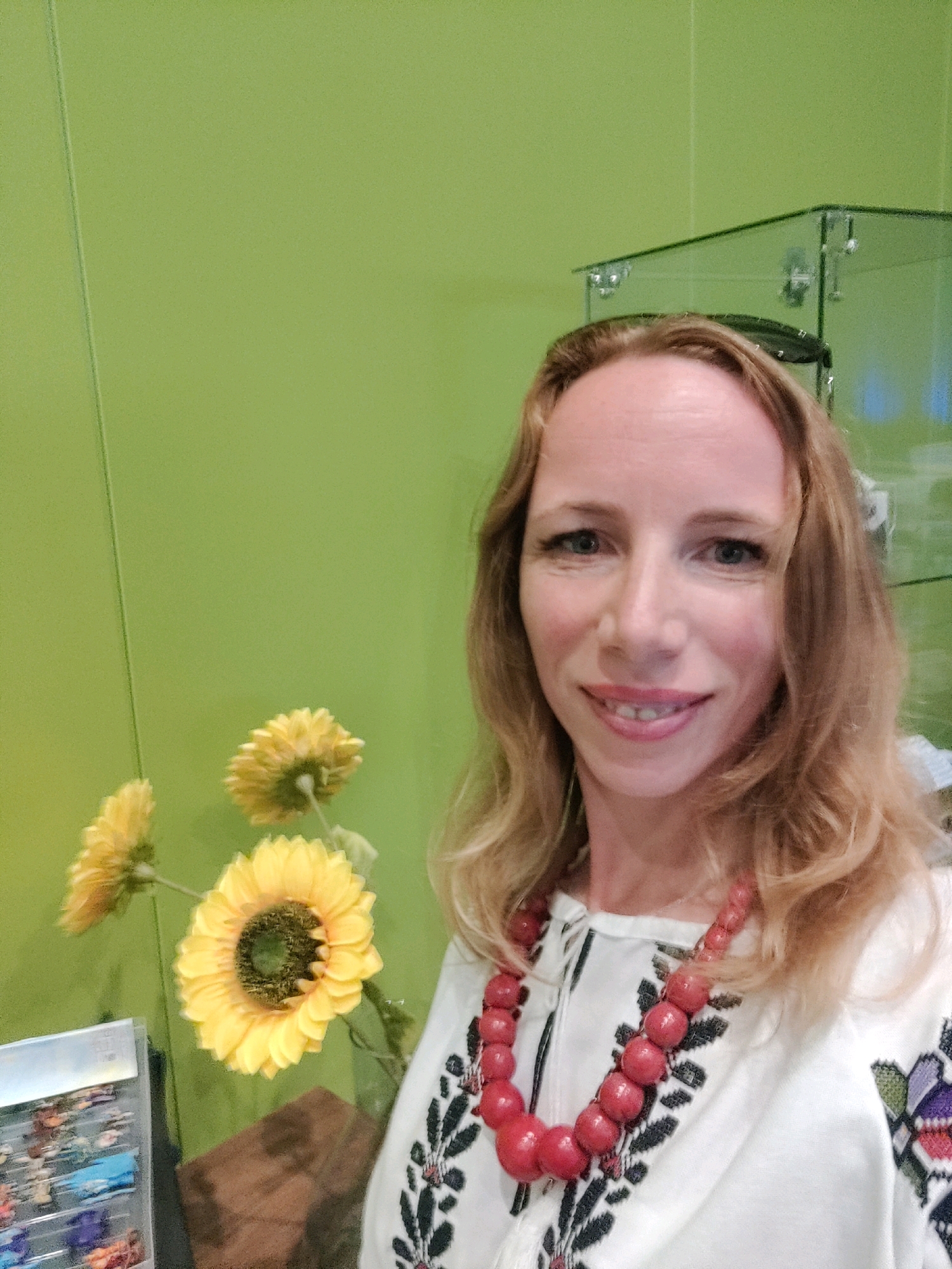 A medium shot shows a woman with long blonde hair and blue eyes smiling at the camera. She is wearing a white blouse with embroidered flowers and a large red beaded necklace. To her left, there are three yellow flowers that appear to be artificial. In the background, a light green wall and a glass display case are visible. The woman is standing in what appears to be a shop or boutique in the city of Groenlo, Netherlands.