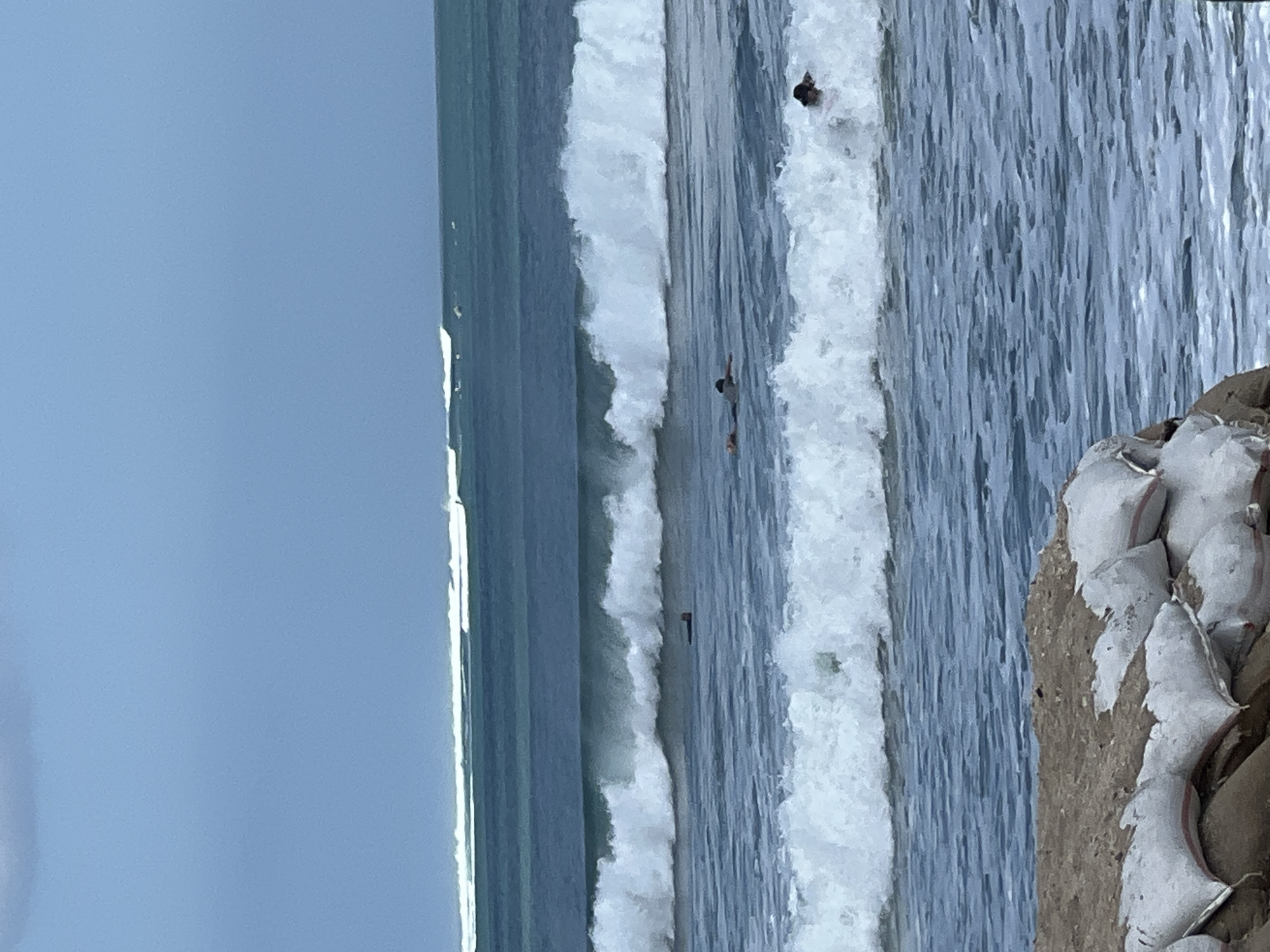 The image depicts a shoreline in Kuta, Indonesia, featuring a beach with sandbags in the foreground, likely for erosion control. The ocean is characterized by multiple rows of waves, with the closest set of waves breaking and frothing white. Further out, larger waves are forming and rolling towards the shore. Several surfers are visible in the water. One surfer is on their board in the middle distance, and another is further out, paddling. Closer to the shore, in the breaking waves, another individual is visible. In the distance, beyond the main set of waves, more surfers and their boards can be seen as small specks on the water. The sky is a pale blue with some overcast clouds.