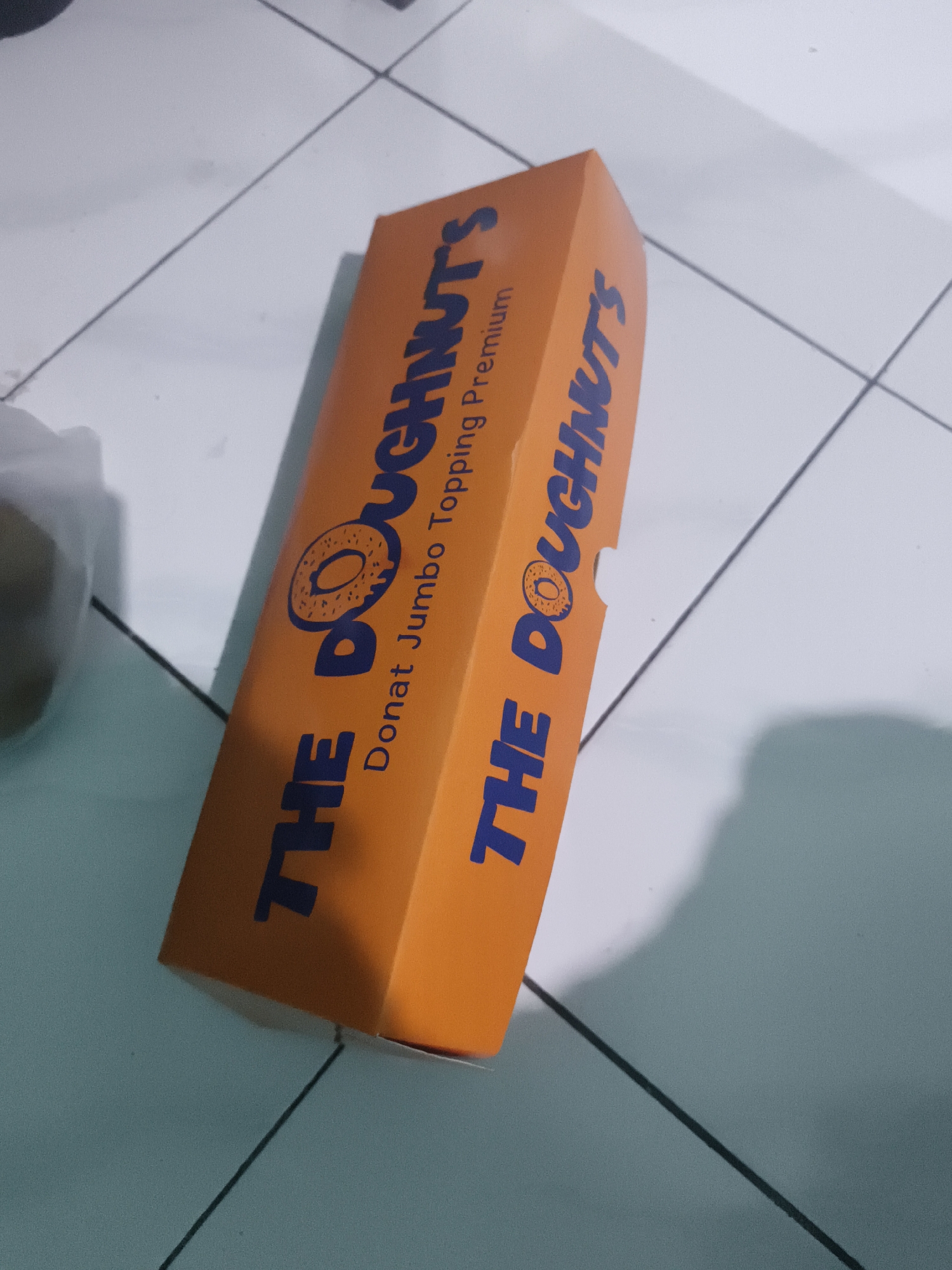 An orange cardboard box, labeled "THE DOUGHNUTS" in blue lettering, lies on a light-colored tiled floor. The box is positioned diagonally, with its right side closer to the viewer and slightly elevated, casting a shadow to the right. The lettering on the box includes the text "Donat Jumbo Topping Premium" below the main title. The tiles are arranged in a grid pattern, with thin dark grout lines. The lighting suggests an indoor environment with a light source from the left. The overall scene is static and focused on the product packaging.