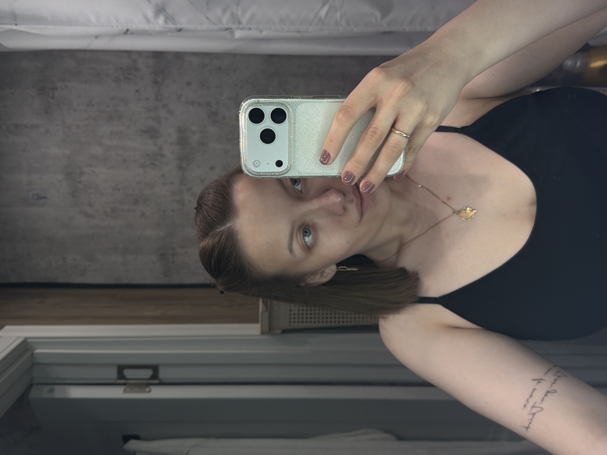 An image, captured as a selfie in a reflective surface, features a woman in an indoor setting, possibly in Reshetnikovo, Russia. The woman, whose head and upper torso are visible, is holding a smartphone with a glittery clear case, covering part of her face. She has brown, possibly chin-length hair, and her blue or green eyes are looking directly towards the camera/mirror. Her nails are painted with a brownish-pink polish, and she wears a simple gold band ring on her right ring finger. A delicate gold necklace with a circular charm, possibly depicting an animal or symbol, is visible around her neck. She is wearing a black, possibly sleeveless, top or dress. On her left upper arm, a tattoo featuring script or handwritten lettering is visible, though the text itself is unreadable.
The background, reflected in the surface, shows a textured grey wall on the left, a white and grey patterned fabric (likely a bed sheet or duvet) at the top, and a wooden horizontal strip below the grey wall. A white window frame or similar architectural detail is visible in the lower left portion of the image. The lighting suggests an indoor setting during daytime or a well-lit interior. The overall impression is a casual, personal photo.