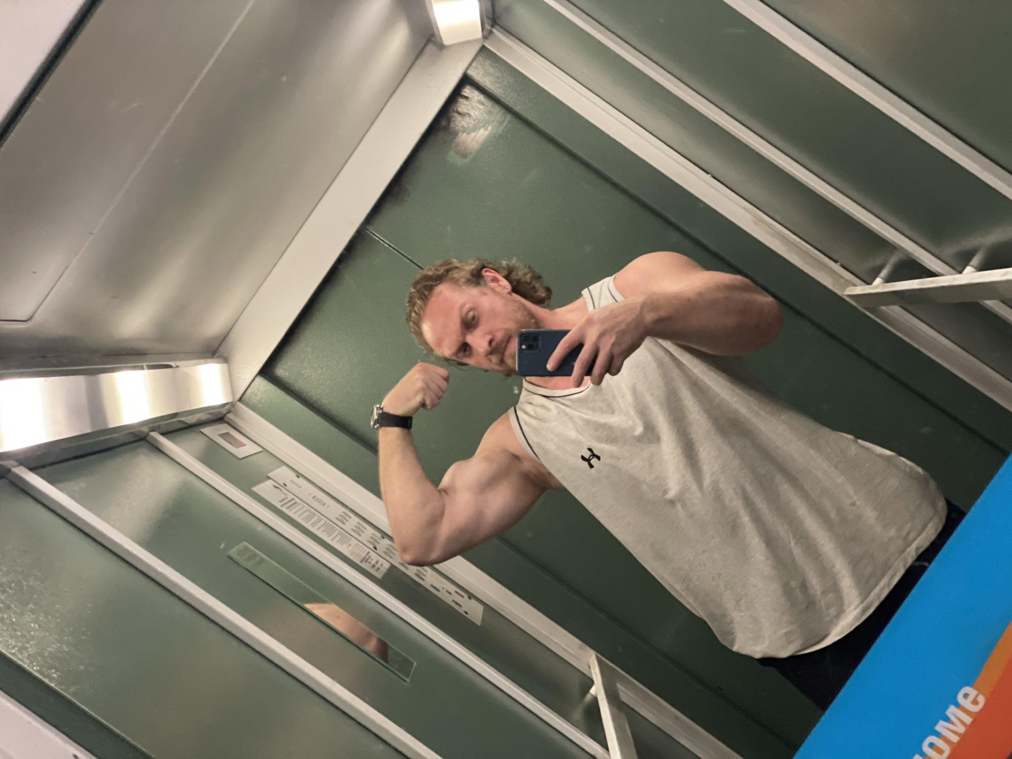 An adult Caucasian male with light skin, brown curly hair, and a beard is standing in an elevator and taking a mirror selfie. He is wearing a light gray tank top with a black logo and black pants. He is flexing his left bicep. He is holding a smartphone in his right hand. The elevator walls are dark green and appear to be paneled with metal trim. There is a mirror on the back wall of the elevator. A portion of a blue and orange sign with white text that reads "OME" is visible in the bottom left corner of the image. The elevator appears to be located in an apartment building in Saint Petersburg, Russia, specifically at the address: St Isaac's Square, building 11, entrance 4, apartment 45.