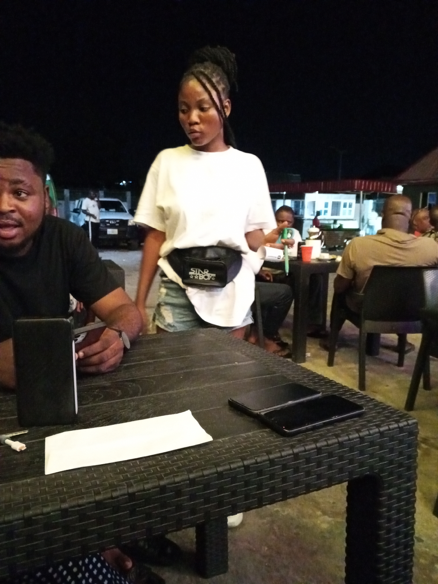 A nighttime scene in Lokoja, Nigeria, shows a group of people at an outdoor establishment. A man in a black shirt sits on the left side of a black woven table, holding a cell phone and looking towards the camera. A white paper wrapper and another cell phone are also on the table. A woman with braided hair, wearing a white t-shirt, denim shorts, and a fanny pack, stands behind the table. Behind her, a young boy sits at another table with a red cup. Further back, another man in a brown shirt is seated. A white vehicle and building are visible in the background.