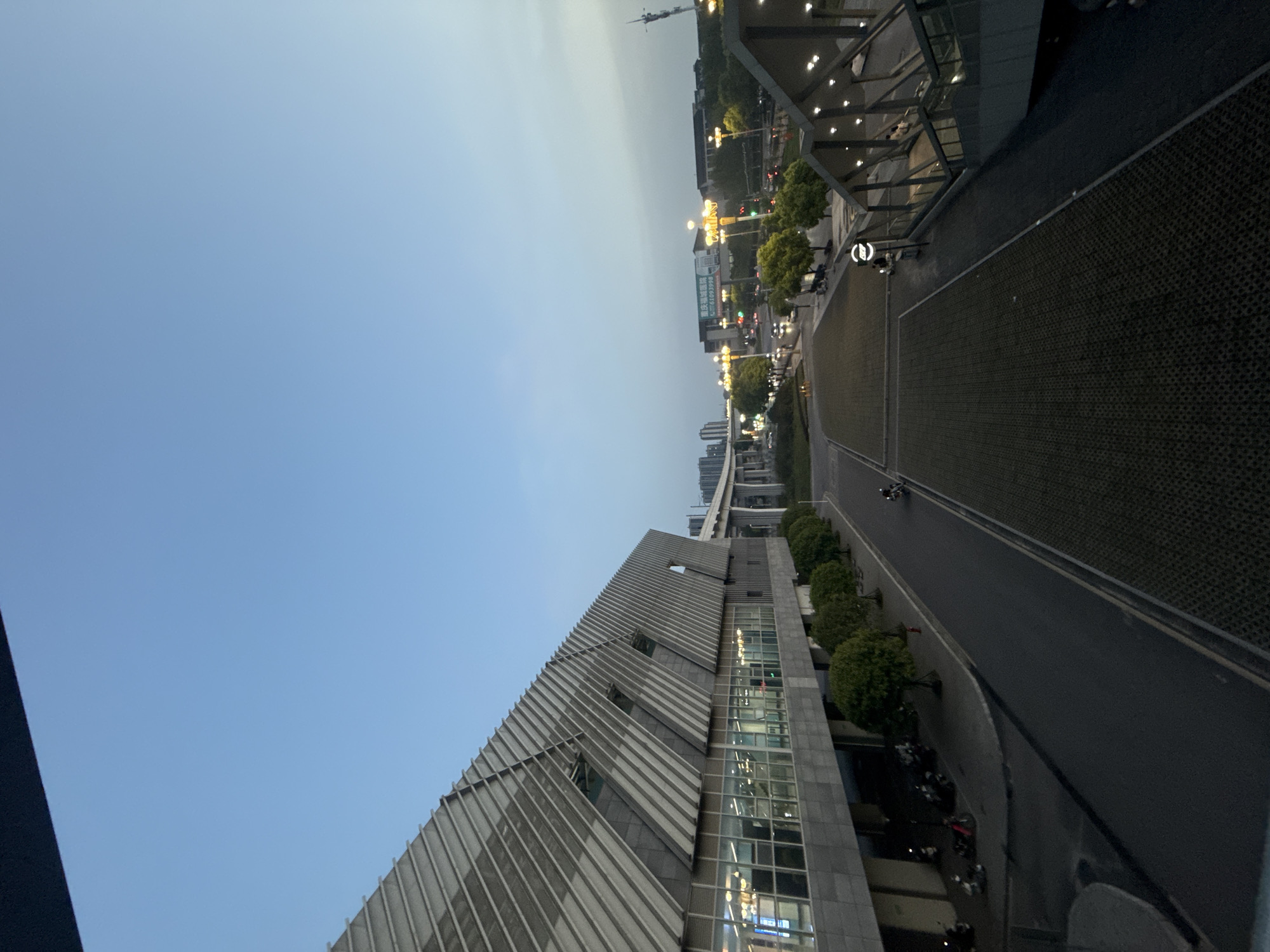 The image shows a cityscape from an elevated perspective in Jiangjin, China. A modern, multi-story building with a distinctive sloped facade and large windows occupies the lower portion of the frame. A paved road runs adjacent to the building, lined with small trees and parked motorcycles. In the background, the scene extends into a city area with other buildings, street lights, and moving vehicles. The sky is light blue, suggesting twilight or early evening.
