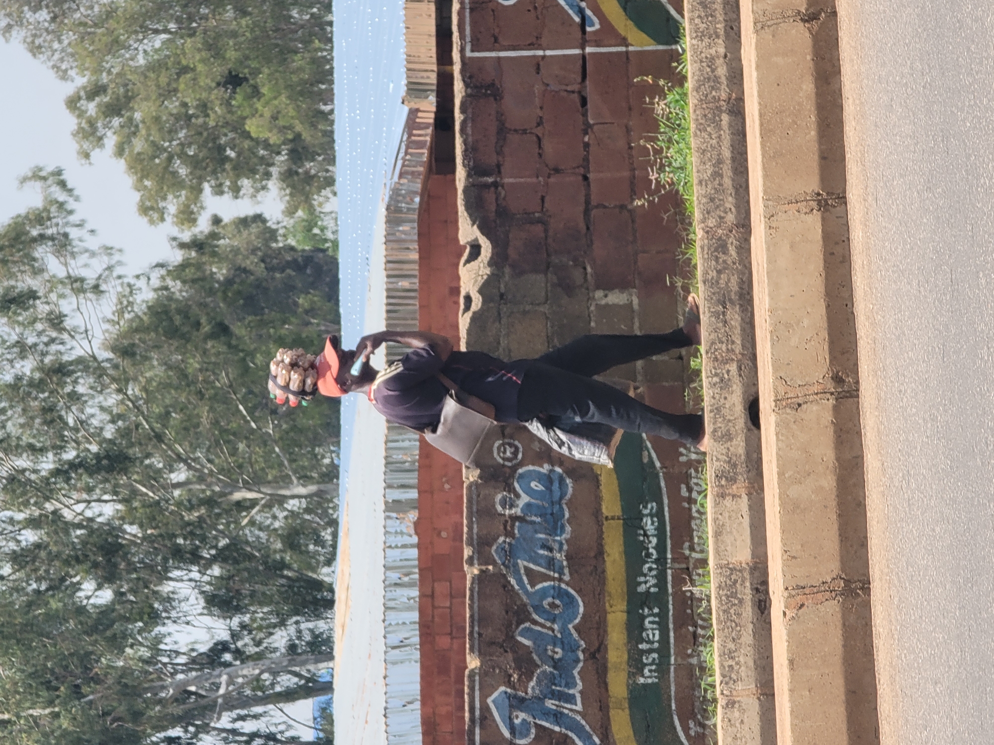 A person wearing a red cap, dark shirt, and dark pants walks along a paved sidewalk next to a brick wall. The person has a dark-colored shoulder bag and is carrying a bundle of items on their head, secured by what appears to be a black band. The brick wall features a mural with blue lettering that reads "IndoMie" and "Instant Noodles" in white text. Behind the wall, there are corrugated metal structures and trees. The sidewalk is bordered by a concrete curb, and the street is visible in the foreground. The sky is overcast. The scene is set in Barakin Lerawi, Nigeria.