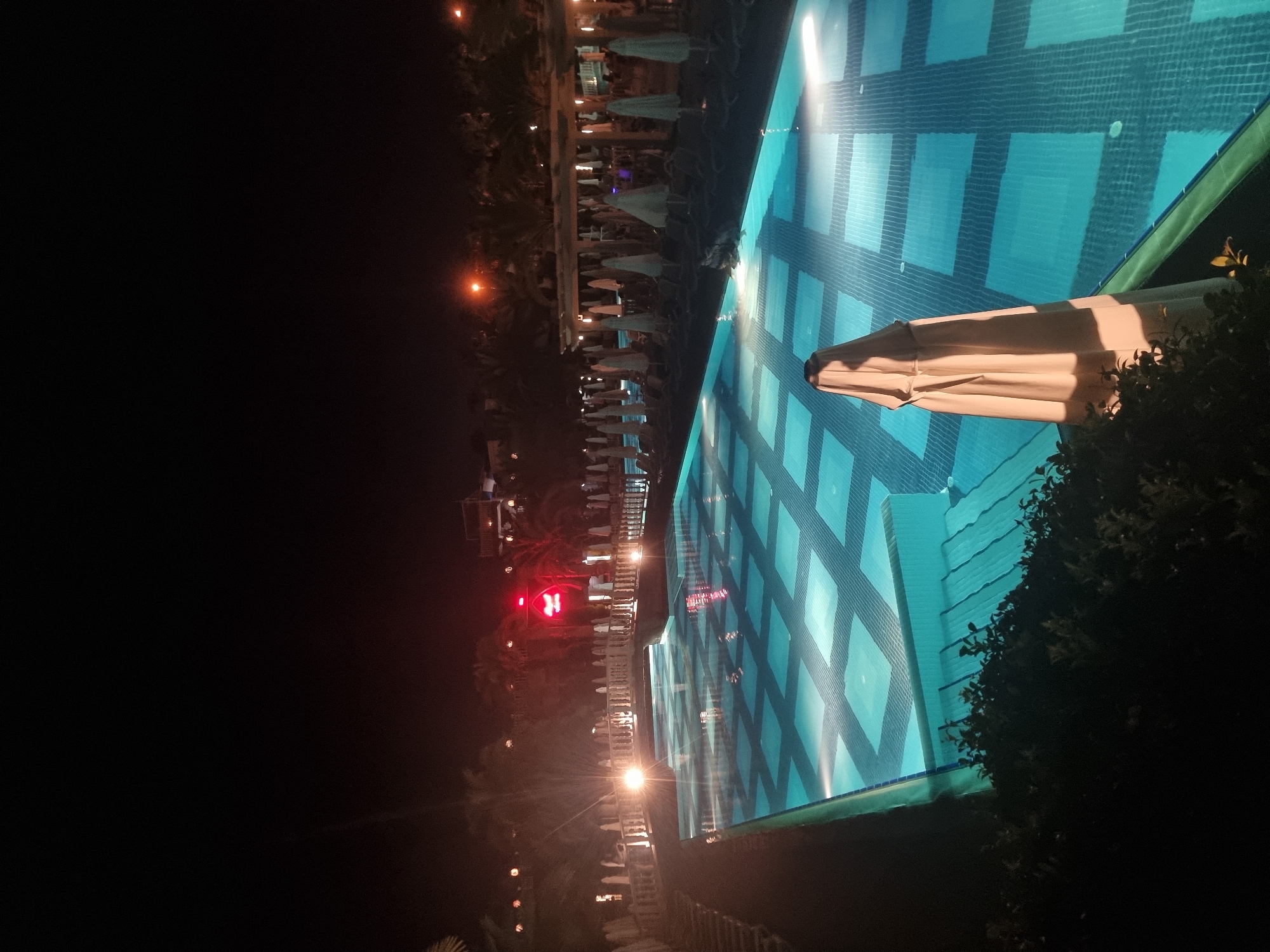 A nighttime outdoor scene features a swimming pool illuminated from below with a geometric pattern of blue lights. The pool is surrounded by lounge chairs and umbrellas, some of which are closed and covered. In the background, there are palm trees and buildings with lights. A red neon sign is visible in the distance. The overall impression is that of a resort or hotel at night. The exact location in Manavgat, Turkey, is not discernible from the image itself.