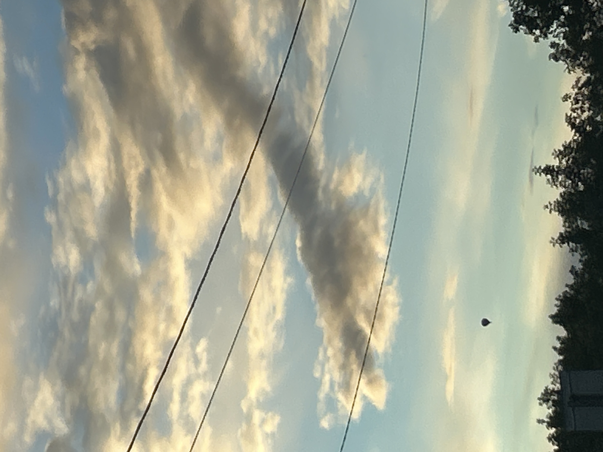 The image shows a wide view of the sky above Yakhroma, Russia, with power lines stretching across the frame. A hot air balloon is visible in the distance, floating against a backdrop of clouds that are illuminated by what appears to be a setting sun, casting golden and grey tones across the sky. The lower portion of the image features a dark silhouette of trees and what might be a building or structure. The scene depicts an outdoor environment, possibly a suburban or rural area on the outskirts of Yakhroma, under a cloud-filled sky with a single hot air balloon.