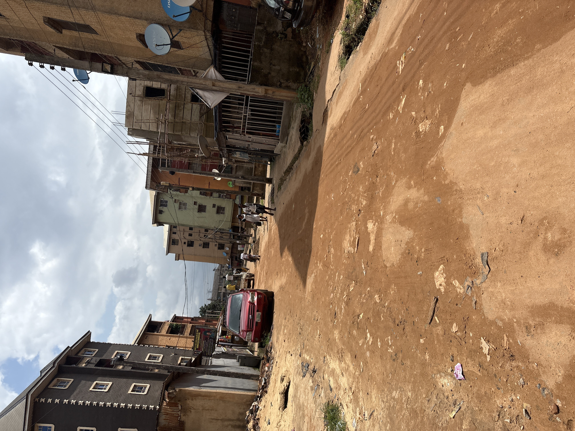 The image depicts a dirt road in the city of Onitsha, Nigeria, with multi-story buildings lining both sides. The road surface is predominantly red earth, showing signs of wear and tear with scattered debris. A red SUV is parked on the left side of the road, facing away from the camera. Several individuals are visible walking along the road and sidewalks.
On the left, a dark gray building with white-framed windows stands prominently. Adjacent to it is a building with orange and brown facades. Further down the street, more buildings of varying heights and colors, including green and yellow, are visible. Some of these buildings appear to be under construction or renovation, indicated by scaffolding.
On the right side of the road, a beige building features satellite dishes mounted on its exterior. A metal-grated enclosure is attached to this building, and beyond it, another dark vehicle is partially visible. Power lines are strung overhead, crisscrossing the scene.
The overall environment suggests a bustling urban area with a mix of residential and commercial structures, characteristic of many Nigerian cities. The condition of the road and the presence of debris indicate potential infrastructure challenges. The sky is partly cloudy, suggesting daytime.