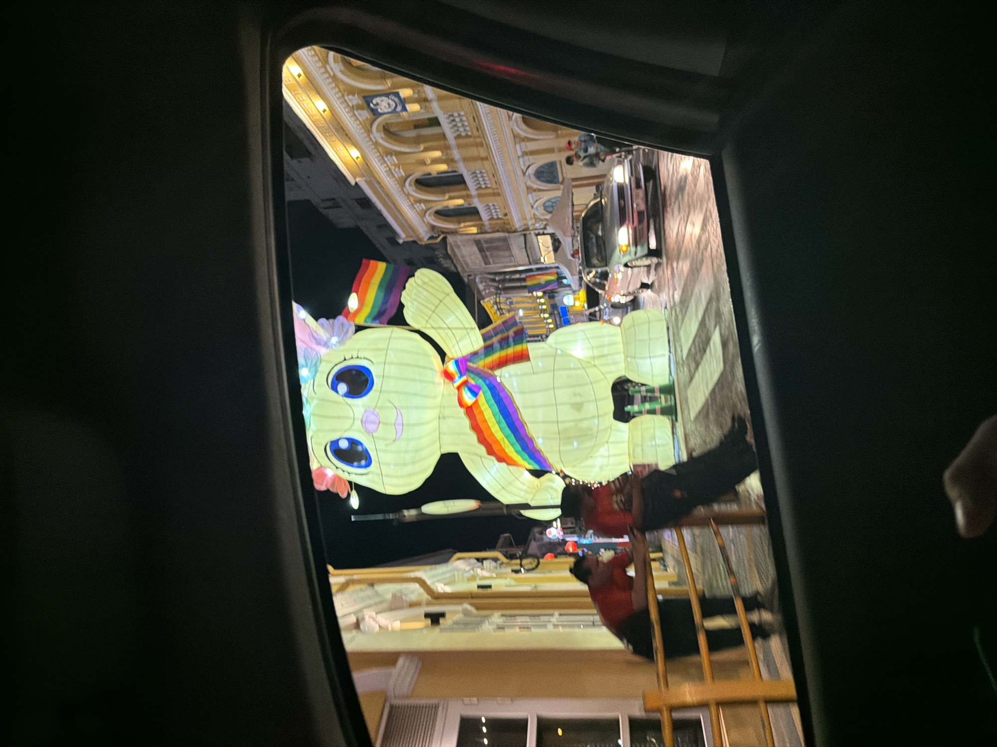 The image, taken at night through a car window, depicts a street scene in Phuket City Municipality, Thailand. Dominating the view is a large, illuminated, cartoon-like rabbit lantern, adorned with a rainbow sash and holding rainbow flags. The lantern is positioned on a pedestrian crossing. Two individuals in red shirts and dark pants are visible leaning on a yellow railing to the left of the lantern. Behind them, the street is lined with brightly lit buildings with arched windows, characteristic of old Phuket Town. A silver car is driving on the street in front of the lantern. The overall atmosphere suggests a festive or celebratory event, possibly related to Pride.