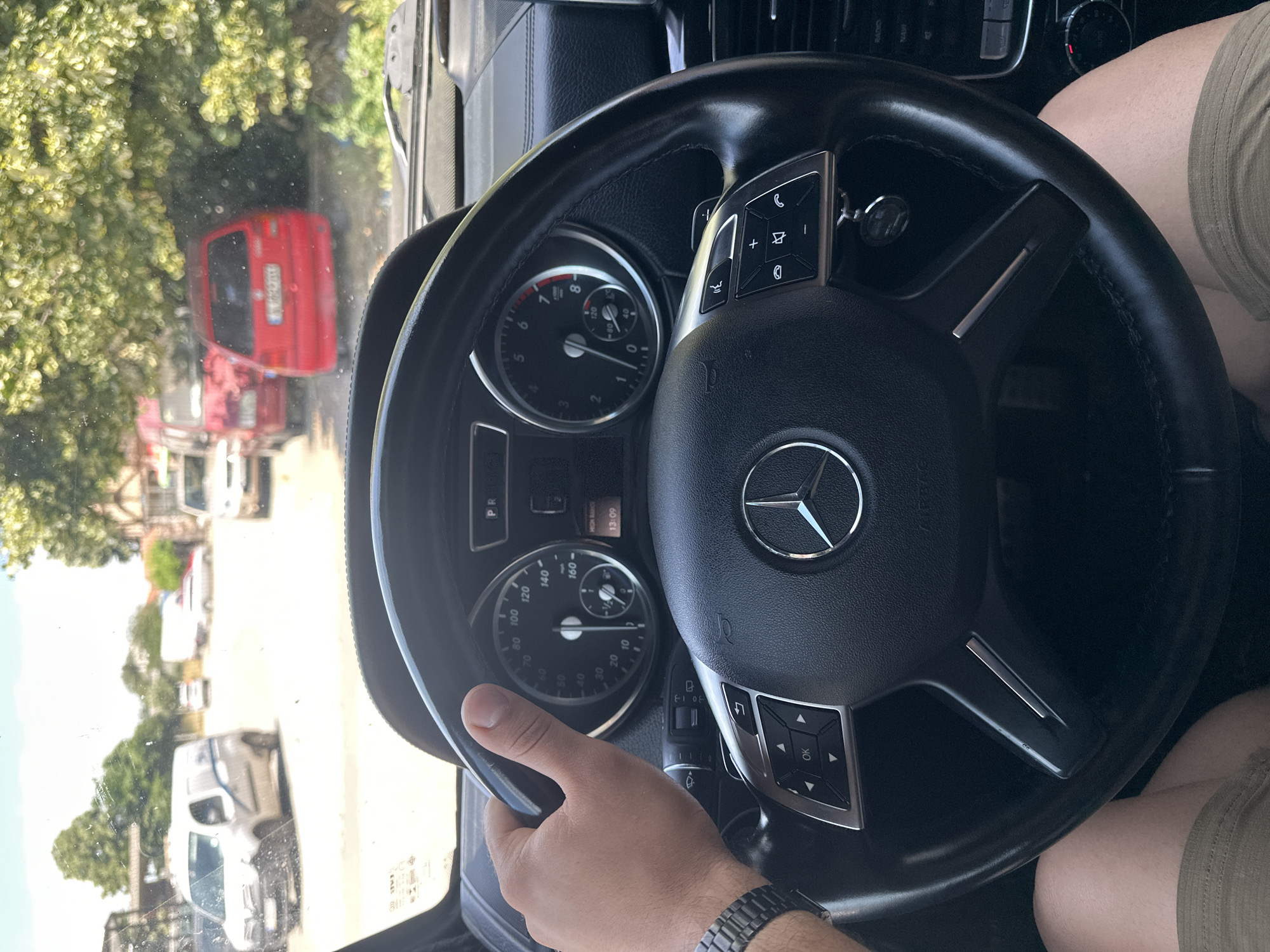 The image shows a first-person perspective of someone driving a Mercedes-Benz G-Class. The driver's left hand is on the steering wheel, and a watch is visible on their wrist. The dashboard features a Mercedes-Benz logo on the steering wheel, a speedometer, a tachometer, and various controls. Through the windshield, several vehicles are visible on a street, including a white van, a red hatchback, and other cars. Trees and buildings line the street, suggesting an urban environment. The license plate on the red car is obscured, but the surrounding scenery and the presence of a local registration plate hint at a location in Bulgaria. Specifically, the visible architecture and the commonality of these car models align with a street scene in Burgas, Bulgaria. The time displayed on the dashboard is 13:09, indicating the photograph was taken in the early afternoon.