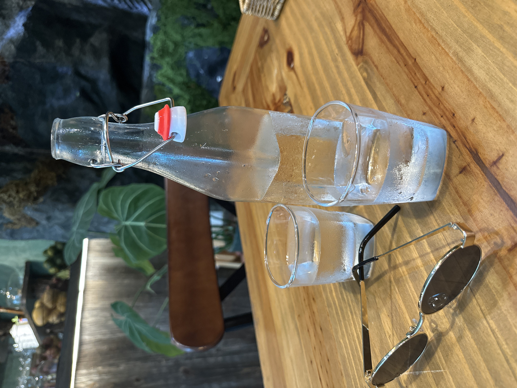 A close-up, horizontally oriented shot captures a casual table setting, featuring a glass bottle, two tumblers, and sunglasses, against a softly blurred natural background.
In the foreground, a clear glass bottle with a swing-top stopper, distinguished by a red and white gasket and metal clasp, lies on its side. Condensation covers the bottle's surface, indicating a cold content. Beside it, two clear glass tumblers are visible; one contains ice and shows condensation, while the other appears empty or nearly so. A pair of stylish sunglasses, featuring circular dark lenses and slender gold frames, rests on the table next to the glasses.
The objects are arranged on a light-colored wooden table with distinct grain patterns. The background is softly blurred, revealing lush green foliage, possibly large-leafed plants, and dark, textured surfaces that resemble rock or stone. A brown, vertical element, possibly a table leg or part of a chair, is also visible in the mid-ground.
The scene suggests a relaxed atmosphere, likely at a cafe or restaurant, where refreshing beverages are served. The presence of sunglasses implies it is daytime, possibly in a warm climate, consistent with the provided location context of Nha Trang Ward, Vietnam. No discernible text is visible within the image.