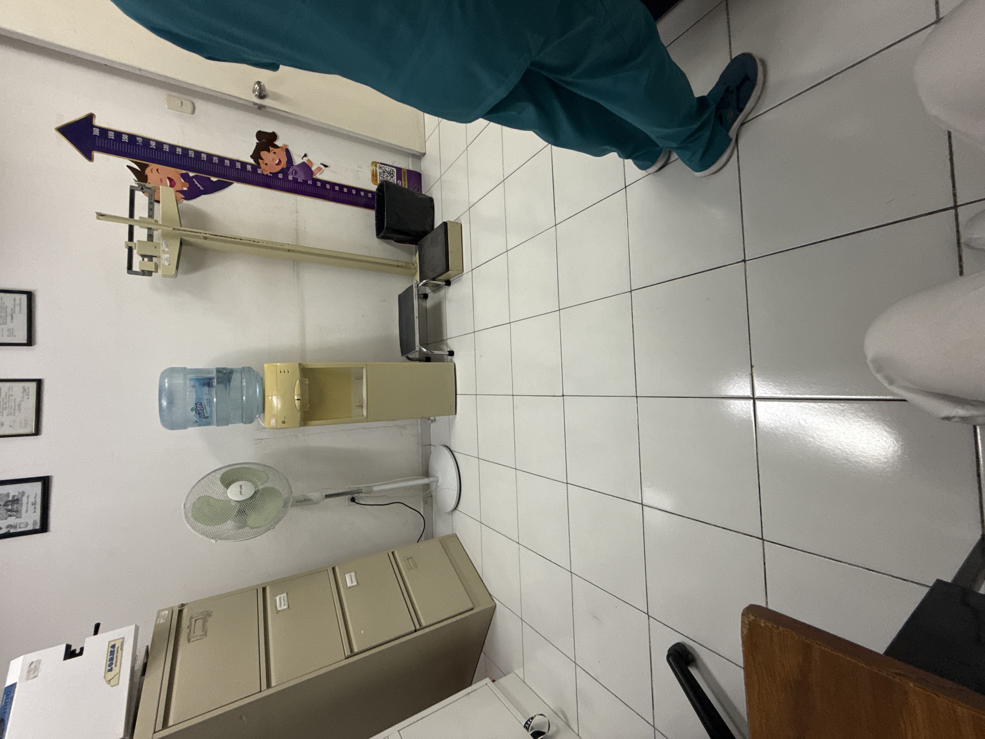 The image shows an interior scene in what appears to be a medical office. The environment is composed of white tiled flooring and white walls. Along one wall, there is a height chart with cartoon figures, medical equipment for measuring height, a medical exam seat, a water cooler, an electric fan, and filing cabinets. Two people are partially visible, both wearing scrubs, which implies a healthcare setting. Based on the image contents, it is likely a clinic or medical examination room located in an institutional setting, with the city details inferred as Puebla, Mexico from external knowledge.
