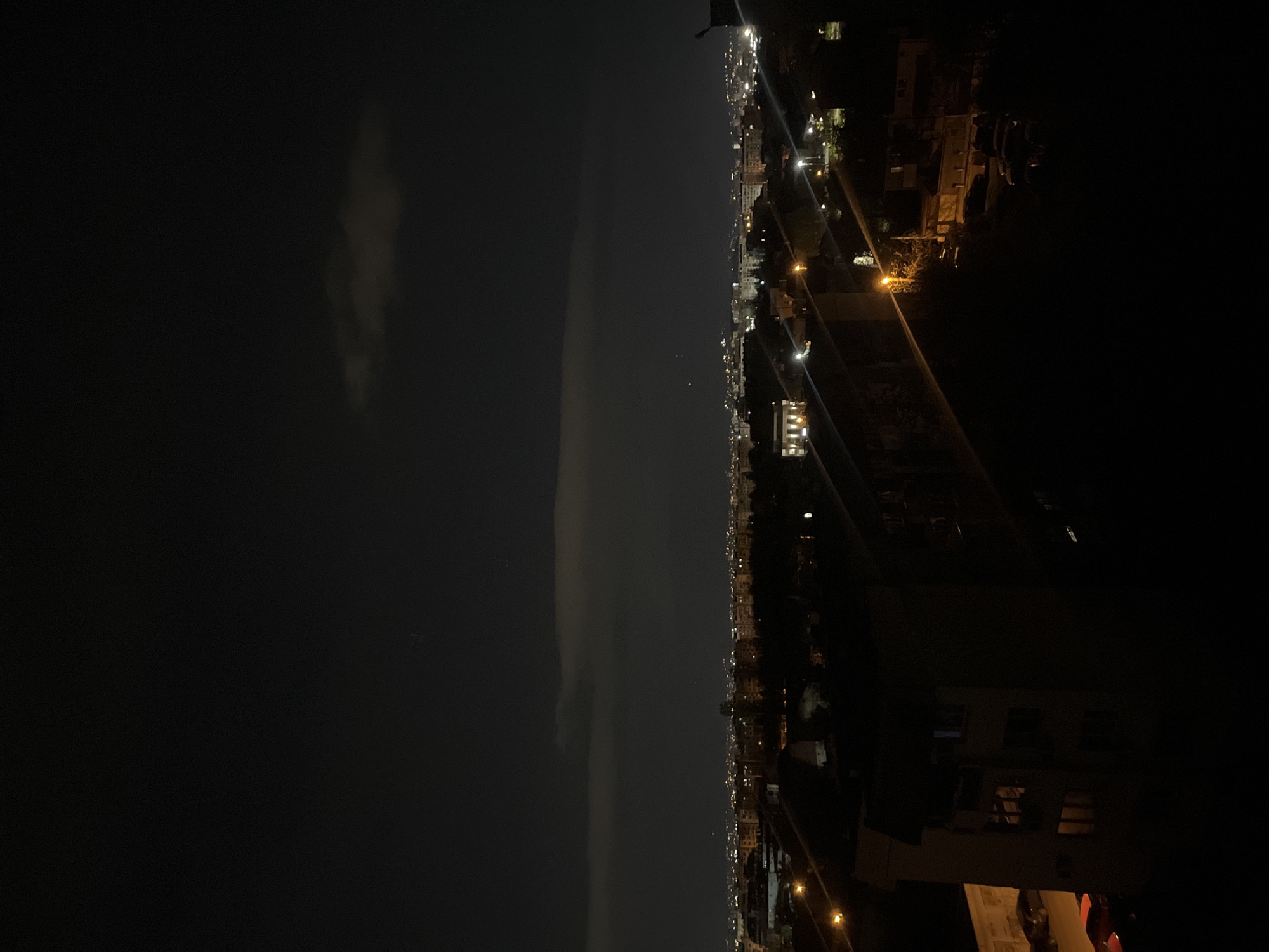 This image captures a panoramic nighttime view of Sofia, Bulgaria, observed from an elevated vantage point, possibly a high-rise building or a hill. The scene is bisected horizontally by the distant urban skyline.
The upper portion of the image depicts the dark night sky, marked by faint, wispy cloud formations or atmospheric haze that create subtle gradients of grey against the black. A few indistinct, tiny points of light, potentially stars or distant atmospheric phenomena, are barely visible.
The lower half of the image showcases the sprawling city, illuminated by a multitude of artificial lights. These lights, varying in intensity and color from yellowish streetlights to brighter white window lights, dot the landscape, outlining buildings and suggesting roads and urban infrastructure. While individual structures are mostly in shadow, the collective glow forms a vibrant network across the cityscape, indicative of a bustling urban environment. A dark, angular railing or balcony structure occupies the immediate foreground on the right, framing the view.
There are no people or specific activities discernible in the image, presenting a static, observational scene. The time of day is clearly night, with no natural light sources other than the extremely faint sky elements. The weather appears to be relatively clear, with some atmospheric diffusion. No visible text is present in the photograph. The overall mood is one of quiet contemplation of an illuminated metropolis at rest.