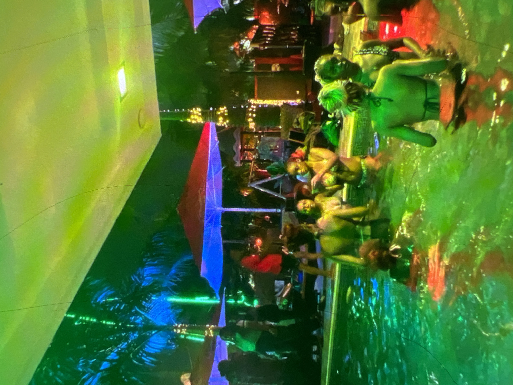 A group of people are gathered in a swimming pool area illuminated by green and yellow lights, suggestive of a nighttime event. Palm trees with blue and green lights wrapped around them are visible in the background, indicating an outdoor setting, likely a resort or a private residence with a pool, common in areas such as Victoria Island or Lekki in Lagos, Nigeria. Several individuals, dressed in swimwear, are standing and interacting within the pool. In the background, there are umbrellas, string lights, and what appears to be a stage or performance area, further suggesting a social gathering or party. The overall atmosphere is festive and vibrant, with the colored lighting creating a dynamic visual effect.