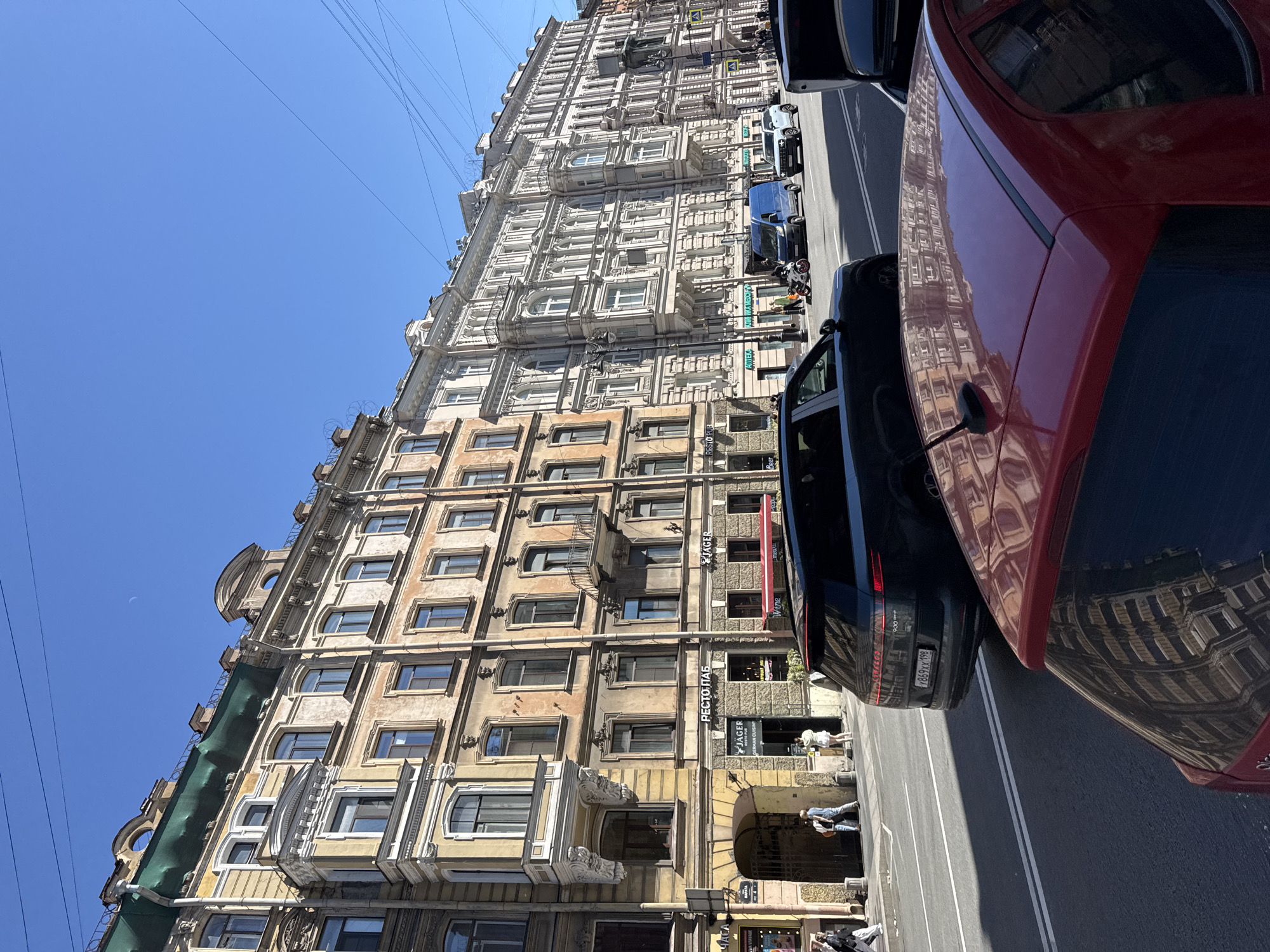 This image captures a sunlit street scene in Saint Petersburg, Russia, likely on Nevsky Prospekt or a similar major avenue, characterized by its grand classical architecture. The foreground features the rear of a red car, its roof reflecting the ornate facades of buildings. To its right, a black SUV is parked, its trunk open. Further down the street, a dark blue minivan and a light-colored SUV are in motion.
The dominant feature of the scene is the row of multi-story buildings lining the street. The building on the left is a warm, faded yellow, adorned with intricate stucco work, balconies, and arched windows. Its lower level features storefronts, including a sign for "РЕСТО-ПАБ" (Resto-Pub) and "JÄGER," with an awning extending over a window. People are seen walking on the sidewalk in front of these establishments.
Adjacent to the yellow building is a second, much lighter-colored building, its facade a pristine white, also showcasing elaborate classical detailing, columns, and balconies. The architecture suggests a historical district of Saint Petersburg. The sky above is a clear, bright blue, with power lines crisscrossing the upper portion of the frame. The overall impression is one of a vibrant urban environment on a clear, sunny day.