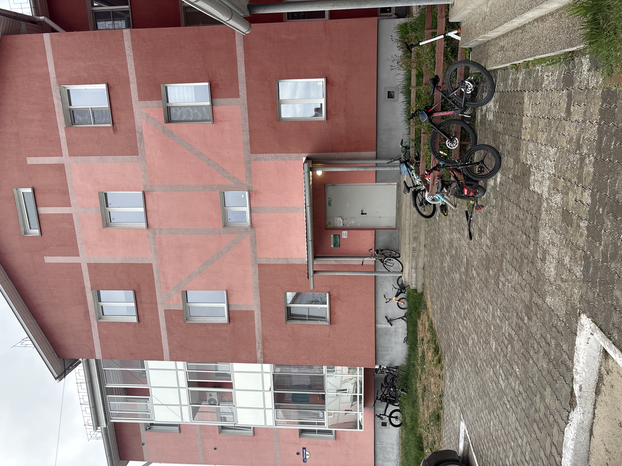The image shows the facade of a red and pink building in Birobidzhan, Russia, with several windows and a gray entrance door. A small sign with Russian text is visible near the entrance. A few bicycles are parked against the building and along a paved walkway. The sky appears overcast. A silver pipe runs horizontally along the top of the building. A modern glass and white structure is attached to the lower levels. A street name sign in Russian is attached to the lower front of the building.
